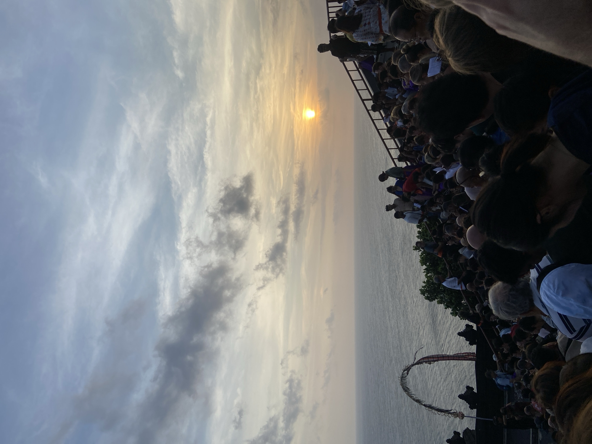 This image depicts a crowd of people gathered on an elevated viewing area overlooking the ocean at sunset. The sun is low on the horizon, casting a warm glow across the sky and water, with scattered clouds adding texture. A railing lines the edge of the viewing platform, and a staircase is visible, with people ascending and descending. In the foreground, the crowd is densely packed, with many individuals looking towards the sea or the sunset.
The scene appears to be at Uluwatu Temple, located in Kuta Selatan, Bali, Indonesia. The distinctive temple architecture is hinted at by shadowy structures at the bottom left, and the cliffside setting is evident from the ocean view. An arched structure, possibly a ceremonial gateway adorned with greenery and fabric, stands near the left edge of the frame. The overall atmosphere suggests a gathering for a cultural performance or to witness the sunset at this renowned location.