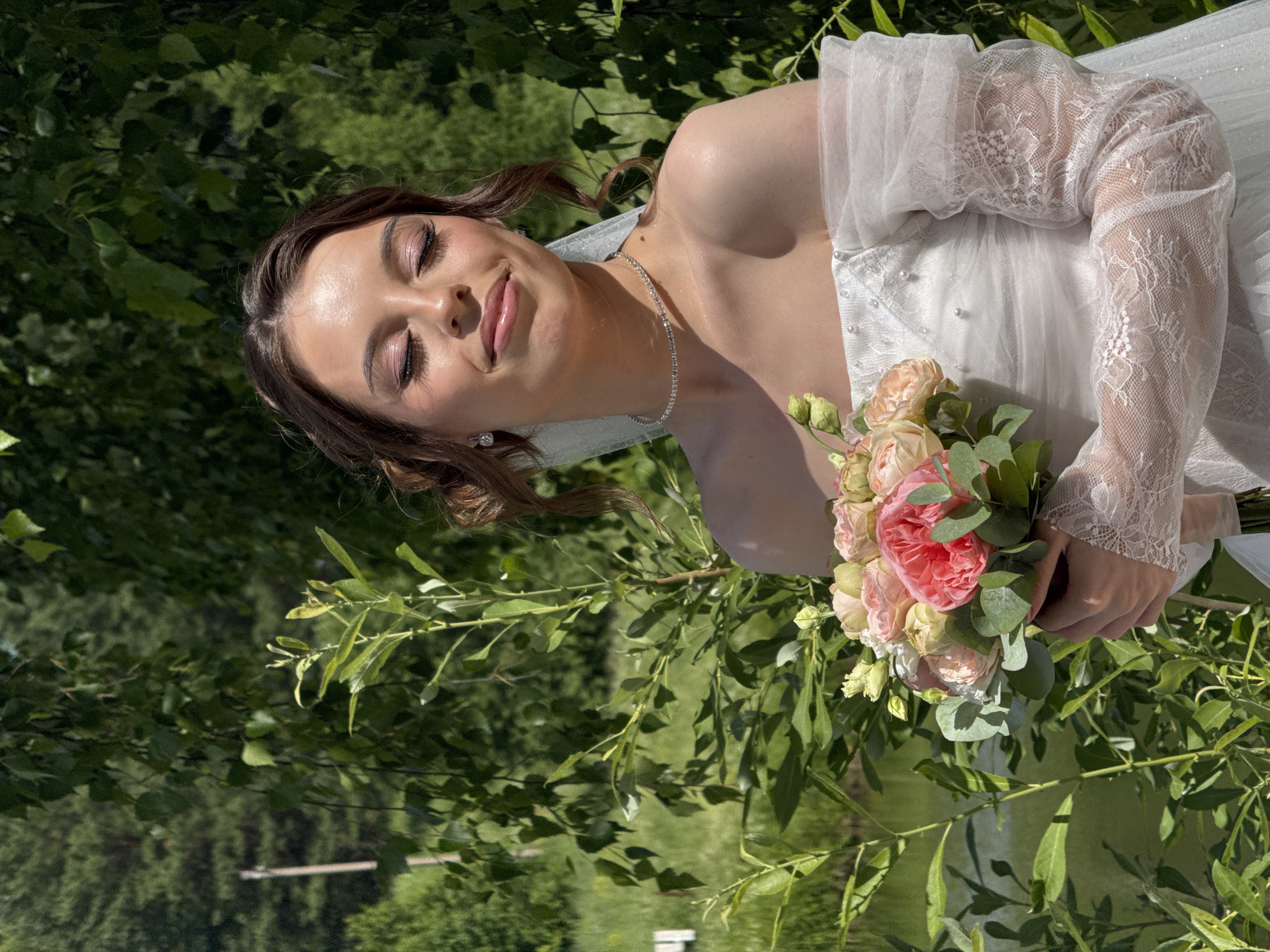 A young Caucasian woman, appearing to be in her late teens or early twenties, is pictured outdoors, possibly in the city of Votkinsk, Russia. She is wearing a white off-the-shoulder wedding dress with delicate lace sleeves and pearl embellishments. She holds a bouquet of light pink and white roses and eucalyptus leaves. Her eyes are closed, and her head is tilted slightly, with a gentle smile on her face. She wears a diamond choker necklace and earrings. The background consists of lush green foliage, with trees and a body of water visible. The lighting suggests it is a sunny day.