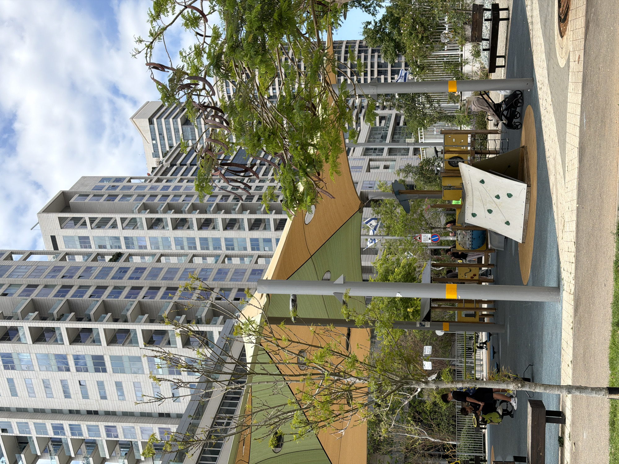 The image captures a scene in Tel Aviv, Israel, specifically a public park or playground area. Dominating the background are tall, modern apartment buildings, characterized by numerous balconies and a repeating geometric pattern. A large, colorful playground structure is centrally located, featuring geometric designs in shades of brown and green. The playground also includes a climbing wall with green holds. Supporting poles, some marked with yellow bands, are positioned throughout the area. In the foreground, trees with sparse foliage add a natural element to the urban setting. A stroller sits near a bench, and a man stands at the bench. Signage, including what appears to be street signs, is visible. A section of sidewalk and a strip of grass border the playground. Flags adorn the tops of the poles along the street. The sky above is partially covered with clouds.