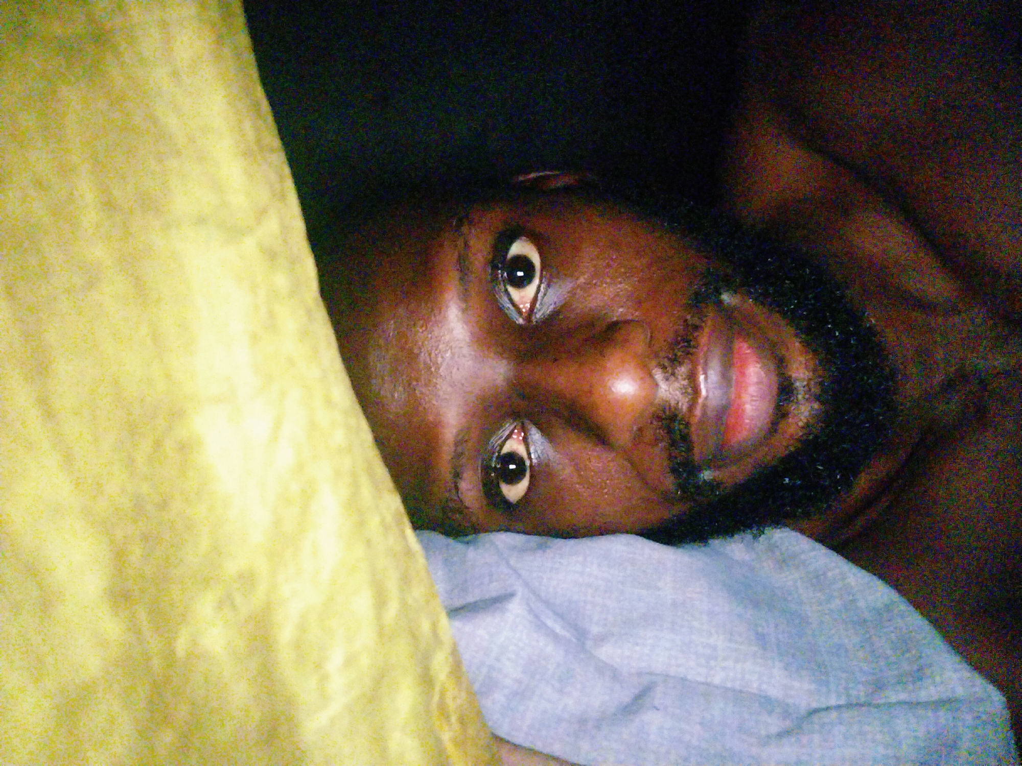 The image is a close-up, tilted, medium shot of a Black individual lying down, with their face oriented towards the camera and positioned on a light blue cloth, likely a pillow or blanket. A portion of a yellow cloth is visible to the left of the individual. The background is dark and indistinct, suggesting an indoor setting. The individual's eyes are open and looking directly at the camera. Their lips are slightly parted, revealing a glimpse of their teeth and the interior of their mouth. The individual has a beard and appears to be resting. The scene does not provide any specific information to determine its location within the city of Uyo, Nigeria.