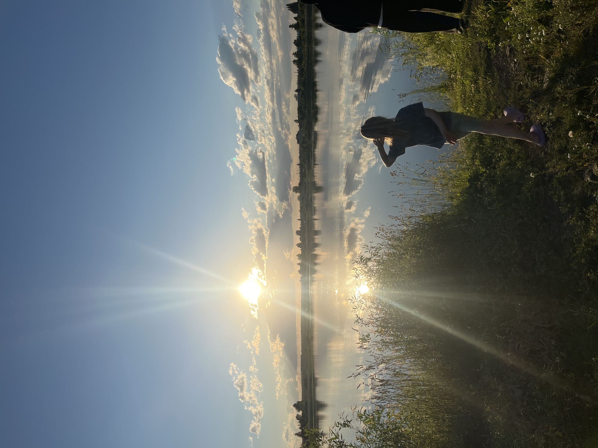 The image depicts a serene natural scene at a lake, likely in or near the settlement of Brusnyatnskoye, Russia, during sunset. The sun is low in the sky, casting a warm, golden light and creating brilliant sunbeams that pierce through the clouds and reflect on the calm water. The sky is a clear blue with scattered, fluffy white clouds tinged with yellow and orange from the setting sun. The lake's surface acts as a mirror, reflecting the sky, clouds, and the distant treeline with remarkable clarity.
In the foreground, on the grassy bank of the lake, are two figures. To the right, the lower half of a person wearing dark pants and what appears to be white socks and black shoes is visible. To the left and slightly closer to the viewer, a young girl is standing, facing the lake. She has blonde hair and is wearing a dark t-shirt and light-colored shorts. Her left hand is raised to her forehead as if shielding her eyes from the sun, and her right hand is behind her back. She is wearing light purple clogs. The bank is covered in green grass and tall, dry reeds, with a few small white wildflowers scattered throughout. The overall atmosphere is peaceful and tranquil, characteristic of a late summer evening.