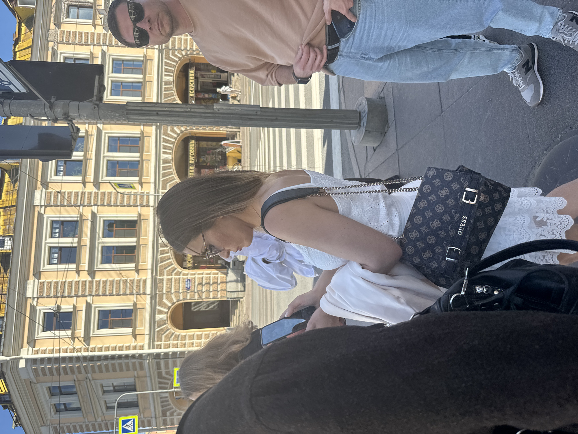 This media file captures a street scene in Saint Petersburg, Russia. The primary subjects are three individuals: a man with sunglasses and a light brown shirt, a woman with a white lace dress and glasses, and another person partially visible in the foreground. The man appears to be standing, looking towards the camera, while the woman is seated or leaning, looking down at her phone.
The scene is set on a sidewalk in front of a building with classical architecture, characterized by ornate window frames and cream-colored stone. A pedestrian crossing with white stripes is visible on the street. The lighting suggests it is daytime, likely sunny, given the shadows and the bright appearance of the building.
Objects of note include a dark patterned crossbody bag with a "GUESS" logo on the woman's shoulder, a black handbag next to her, and a smartphone in her hands. The man has a watch on his left wrist and his phone is tucked into his waistband. A traffic sign with a blue triangular symbol is visible in the lower left corner.
The activity appears to be casual; the individuals seem to be waiting or resting on the sidewalk. The atmosphere is relaxed, with no indication of any particular event. Visible text includes "GUESS" on the bag and Cyrillic lettering on storefronts in the background, with one clearly legible as "ИСКУСНИЦА". The overall impression is a typical urban moment captured on a clear day.