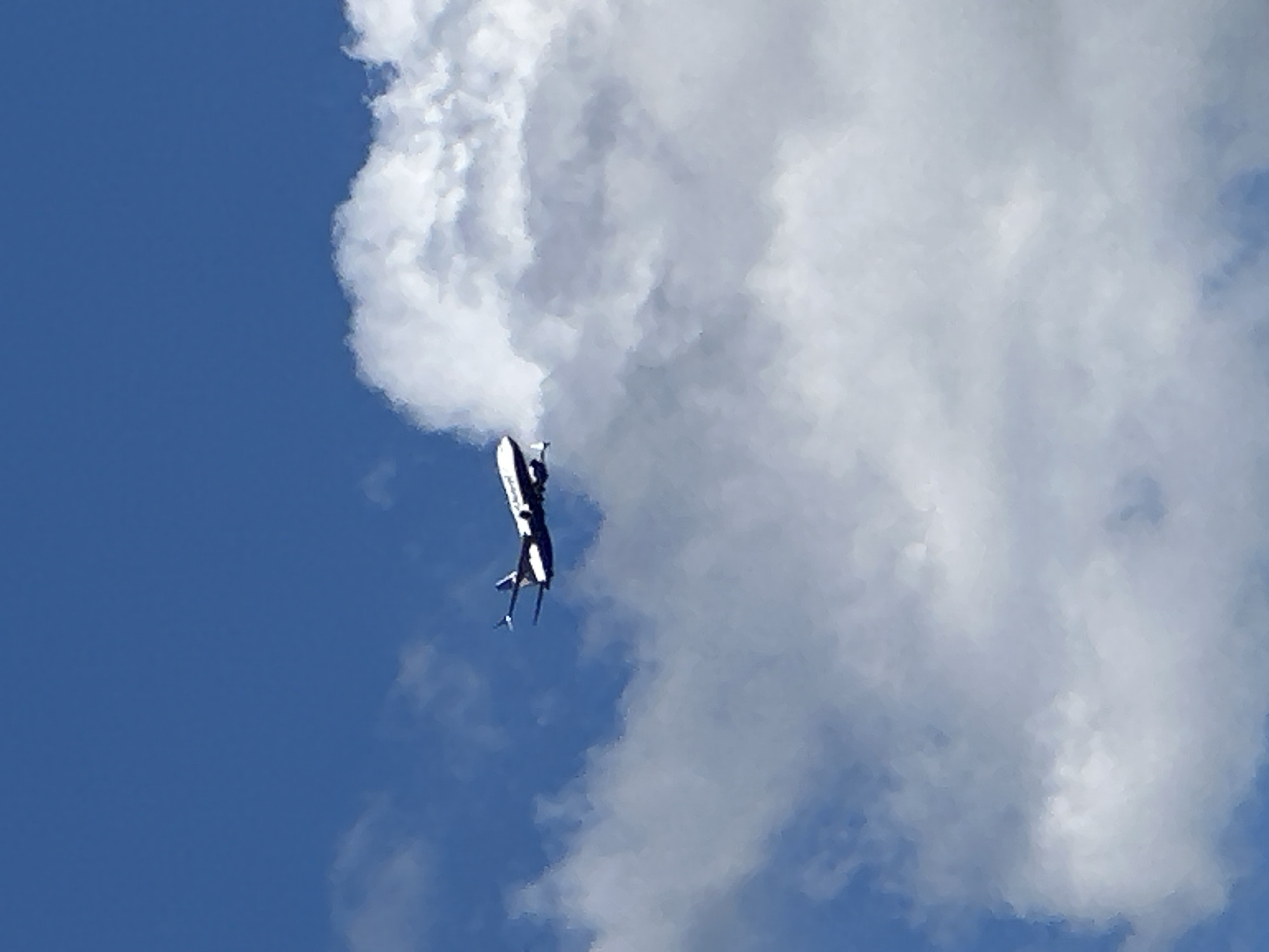 An airplane is seen in mid-flight against a blue sky with scattered clouds. The airplane is dark colored with white accents, and it appears to be a commercial jet. The plane is angled slightly upwards, with its wings extended. The clouds are white and puffy, and they are spread across the sky. The image is taken from below the airplane, looking up.