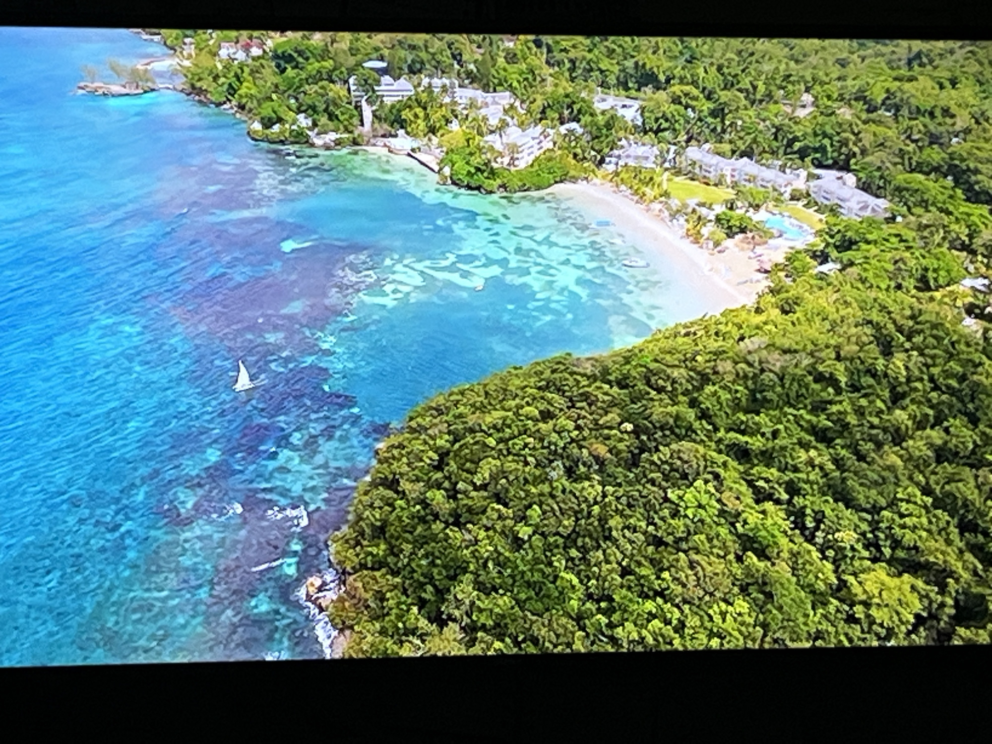 This image is an aerial view of a tropical coastline. The scene features vibrant turquoise waters with visible coral reefs near the shore, transitioning to a deeper blue further out. A small white sailboat is visible on the water. The coastline is characterized by a sandy beach backed by lush, dense green vegetation and trees. Several white buildings, possibly resort accommodations, are nestled within the trees along the coastline. The image does not depict any recognizable landmarks or features associated with the city of Kharkiv, Ukraine.