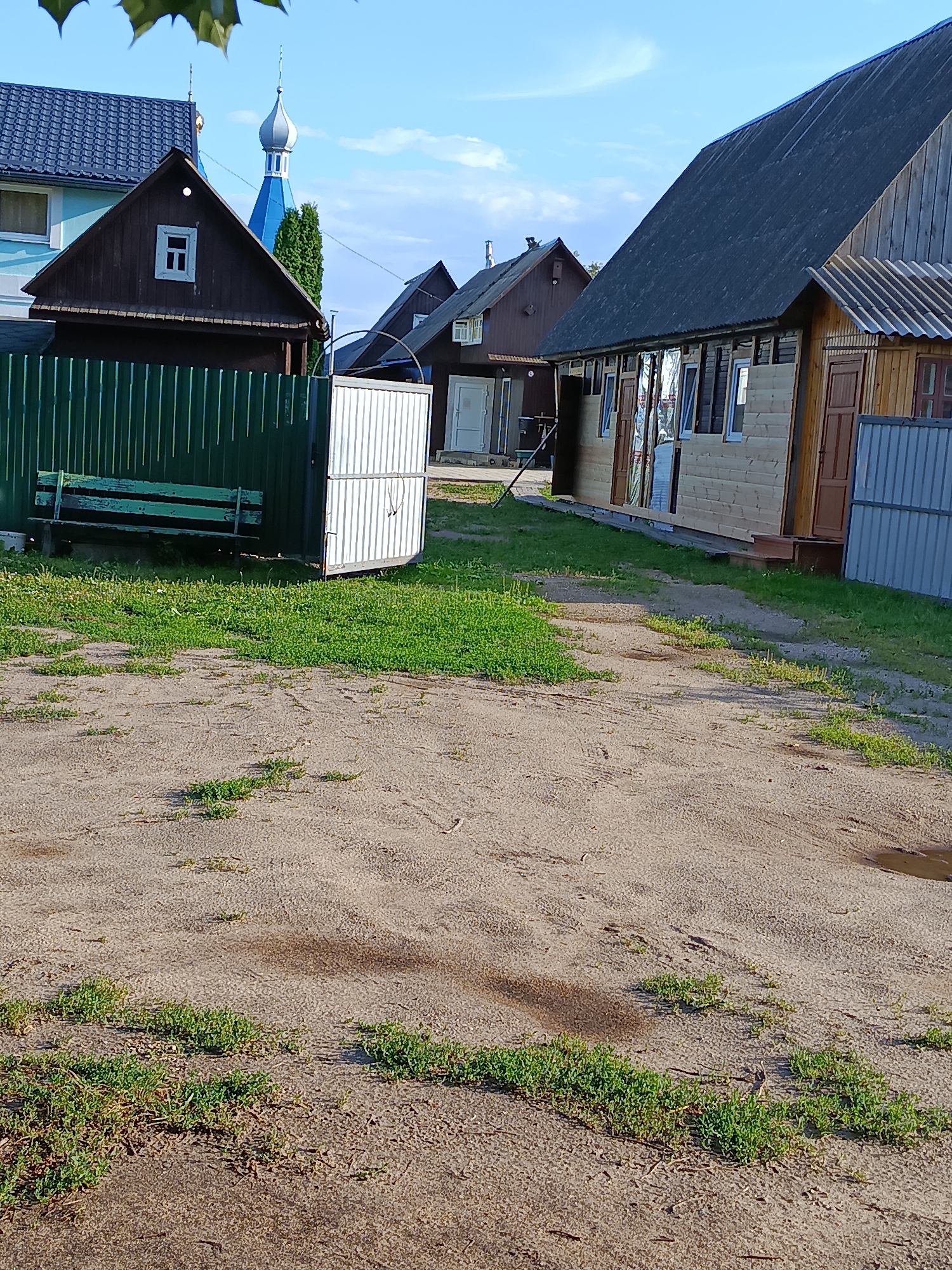 The image depicts a street scene in Smalyavichy District, Minsk Region, Belarus. In the foreground, a sandy path with sparse patches of grass and puddles of water leads towards several buildings. To the left, a dark green metal fence encloses a yard with a weathered green wooden bench. Beyond the fence, a dark brown wooden house with a white-framed window is visible, with a light blue house and a blue-domed building behind it. To the right, a light-colored wooden building with a dark, sloped roof and multiple windows is situated. A light grey metal fence runs along the side of this building. The overall scene suggests a rural or semi-rural residential area under a clear blue sky.