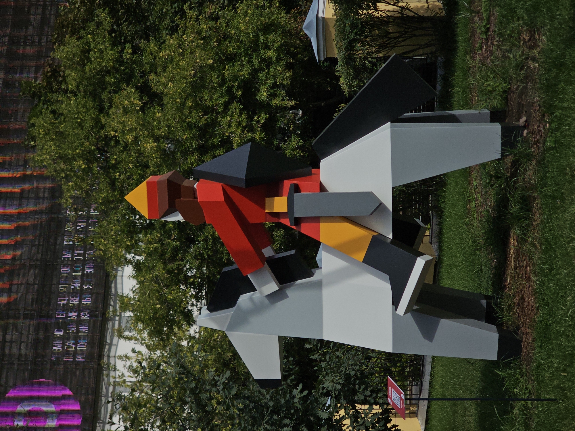 The image shows a stylized, geometric sculpture of a knight on horseback in an outdoor setting. The sculpture is made of flat planes of color, primarily white, gray, red, yellow, and black. The knight wears a red tunic with yellow accents, black leggings, and a yellow and red helmet. The horse is white and gray with black accents. The sculpture is positioned on a grassy area, with trees in the background. A small red sign with a QR code is visible in the foreground. In the background, a large, dark screen displays abstract, colorful patterns and text, possibly a digital billboard or facade. The architecture of a building with a yellow facade and a light blue roof is also visible behind the trees on the right. This sculpture is part of the art installation "Geometric Dreams" by Georgy Frangulyan, located in the Patriarch's Ponds area of Moscow, Russia.