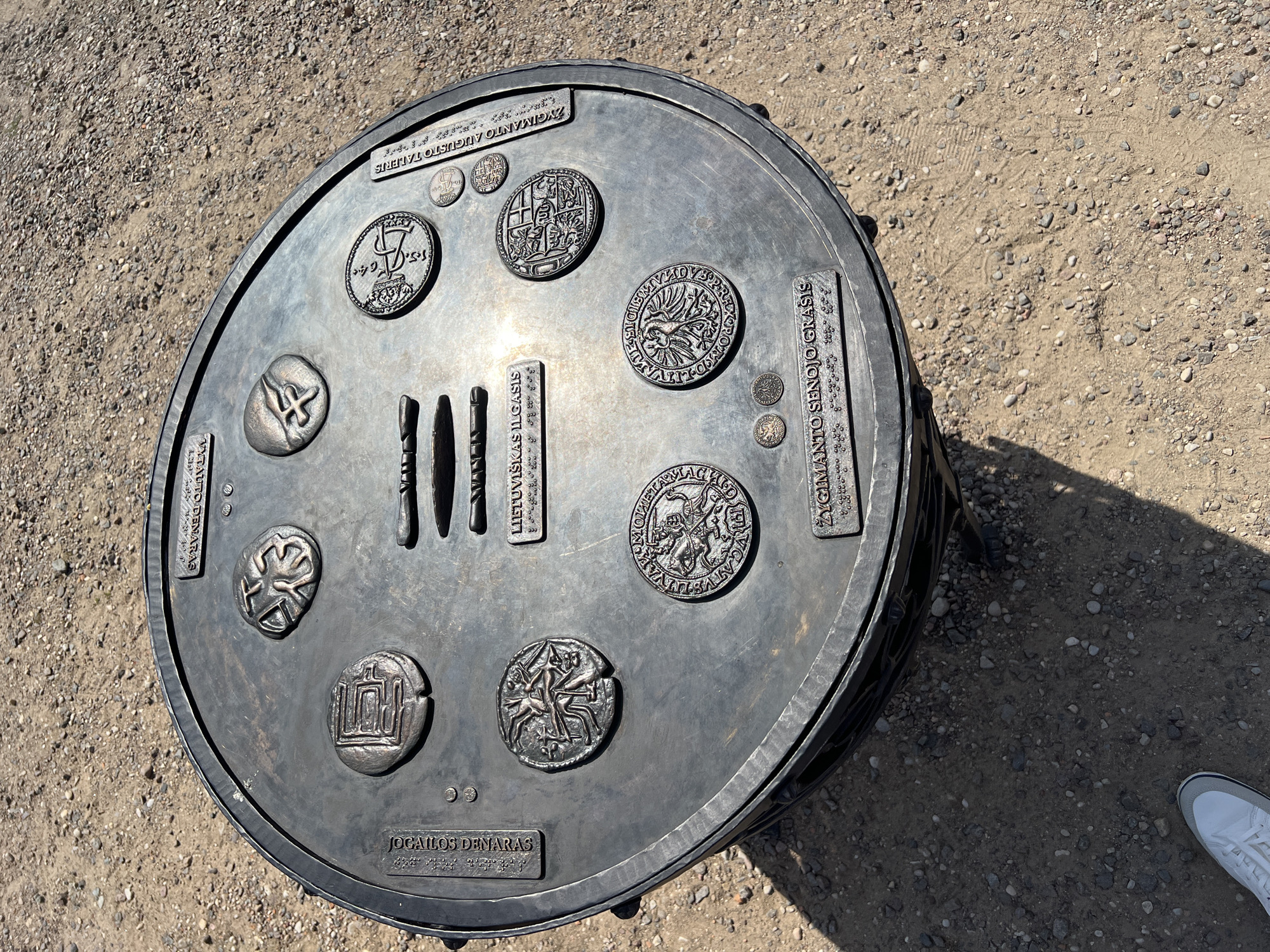 The image shows a circular, bronze monument in the city of Trakai, Lithuania, placed on a gravelly ground. The monument depicts various historical coins and symbols, possibly representing the monetary history of Lithuania. Inscriptions in Lithuanian and braille are visible on the monument's surface. The monument appears to be a public art installation or a historical marker. The background consists of dry, sandy soil with scattered pebbles and some shadows cast by the monument. No people are clearly visible in the image.