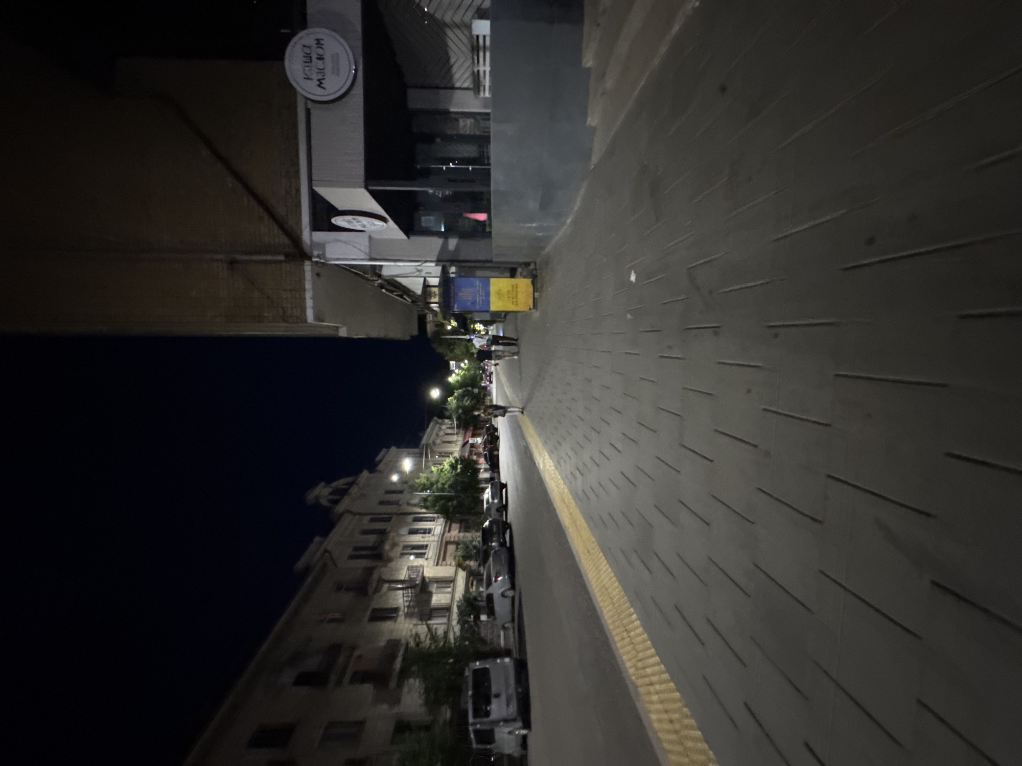 The image captures a nighttime street scene in Chernivtsi, Ukraine, likely on Kotsubynskoho Street given the architecture and the presence of a cafe sign reading "KAWA MACIOR." A sidewalk is prominent in the foreground, paved with dark gray rectangular tiles arranged in a staggered pattern. A textured yellow tactile paving runs along the edge of the sidewalk bordering the street.
On the street, several parked cars are visible, including a silver van on the left, and other cars further down the road. The buildings lining the street are multi-story structures with visible windows and balconies. One building on the right appears to be a cafe or restaurant with a prominent circular sign. A billboard or display board with blue and yellow panels, possibly a Ukrainian flag motif, stands near the cafe entrance.
A few individuals are walking on the sidewalk. One person is walking towards the camera on the right side of the frame, carrying a bag. Further down the street, two people are walking away from the camera. Streetlights illuminate the scene, casting a dim glow on the surroundings. The sky is dark, indicating it is nighttime. The overall atmosphere is calm and urban.