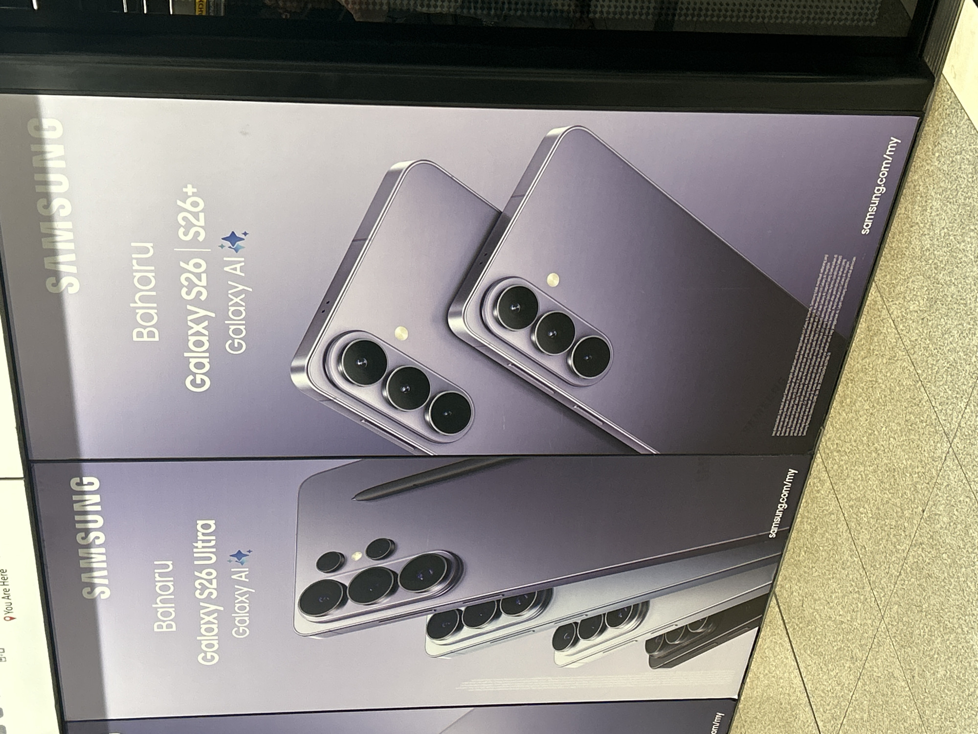 The image shows a billboard advertisement for Samsung Galaxy S26 Ultra and Galaxy S26+ phones, featuring their cameras and the "Galaxy AI" branding. The billboard is in a public space, likely a transit station, as indicated by the tiled floor and the presence of a metallic barrier or wall. The advertisement is displayed in two panels. The left panel features the "Galaxy S26 Ultra" and showcases a progression of phones, while the right panel highlights the "Galaxy S26 | S26+" and shows two phones stacked on top of each other. The text is in Malay. The overall color scheme is purple and silver, reflecting the phones' design. The bottom of the billboard displays "samsung.com/my", indicating a Malaysian website. The presence of the "You Are Here" marker in the upper portion of the original image, albeit partially cut off, and the website domain suggest the location is in Kuala Lumpur, Malaysia.