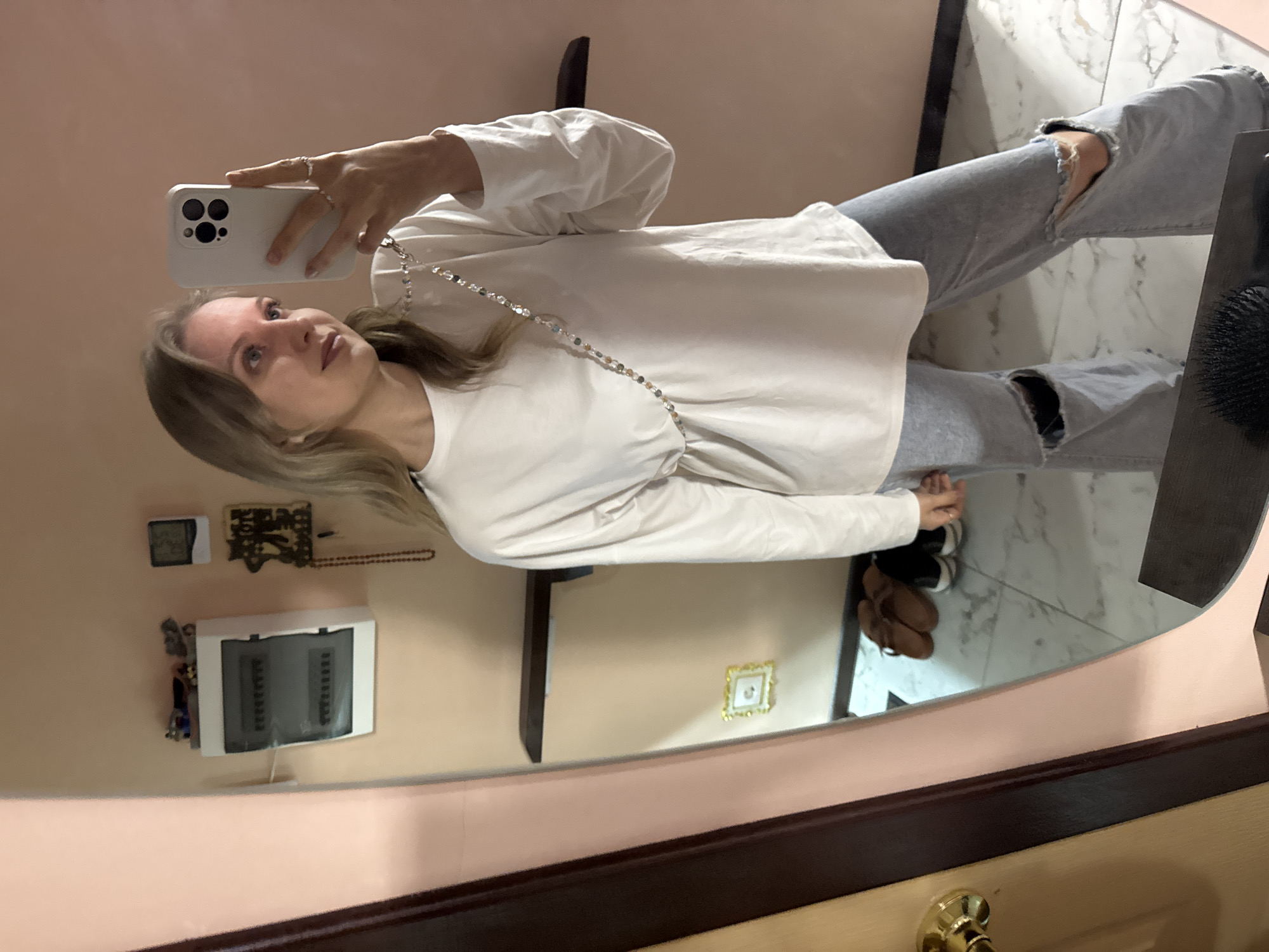 A young woman with blonde hair takes a mirror selfie indoors. She is wearing a white, long-sleeved shirt and light blue jeans with large rips at the knees. A beaded lanyard is draped around her neck. Her left hand is holding a white smartphone, and her right hand, adorned with rings, is resting on her hip. The background shows a pale pink wall with a digital clock, a wall-mounted electrical panel, and a shelf. In the reflection of a large mirror to the left, a doorway with a brass doorknob and a wooden doorframe are visible. Below the shelf in the mirror's reflection, a small framed picture hangs on the wall, and a pair of brown shoes and black sneakers are placed on a marble-tiled floor. The scene appears to be set in a domestic environment, possibly an apartment, in the city of Bolshaya Venya, Russia.