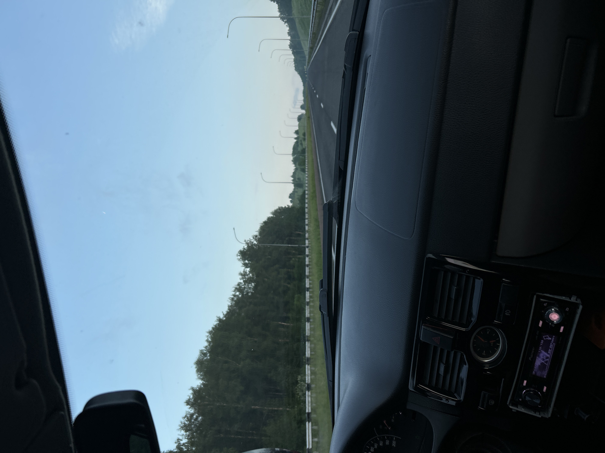 The image shows a view from the inside of a car, looking forward through the windshield. The dashboard is visible in the foreground, with an analog clock and a car stereo. Outside the car, a paved highway stretches into the distance, lined with trees and streetlights. The sky is a clear, pale blue, with a few wispy clouds. The scene appears to be set in a rural or suburban area, possibly during the early morning or late evening. There are no people or animals visible in the image. The overall impression is one of a calm and peaceful drive.