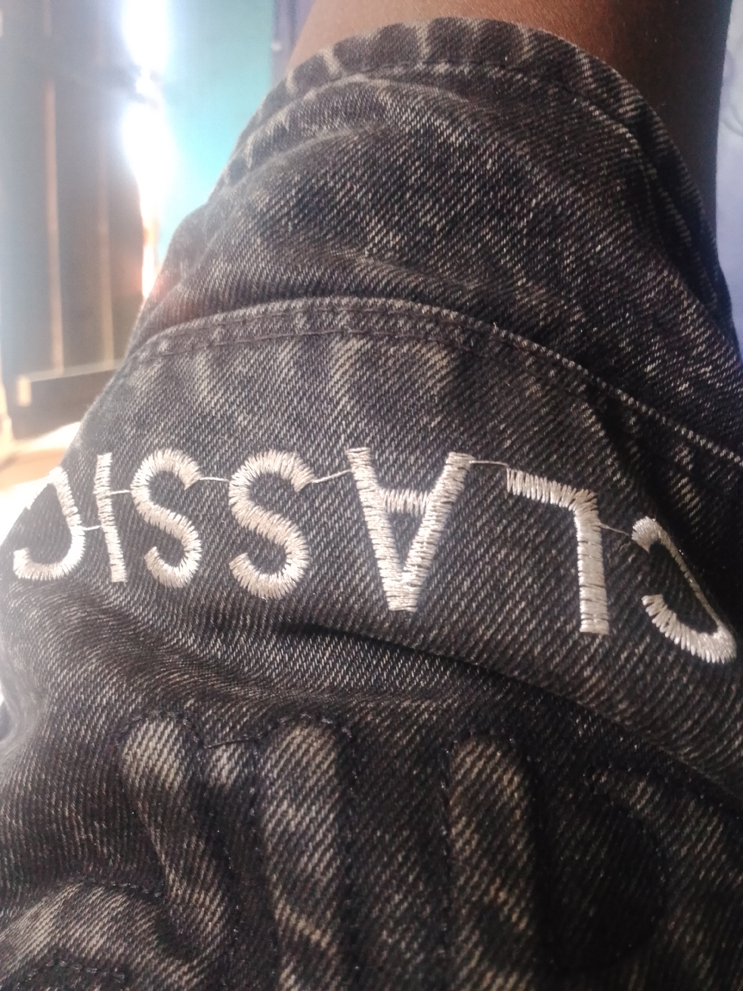 A close-up shot captures the word "CLASSIC" embroidered in white thread on a dark denim fabric. The embroidery is slightly raised and has a textured appearance. The denim material shows a diagonal weave pattern and appears to be worn or faded in some areas. The lighting in the image is bright, creating highlights and shadows on the fabric's surface. The background is blurred, with hints of light and color, suggesting an indoor or outdoor setting. The phrase "CLASSIC" is centered across the image.