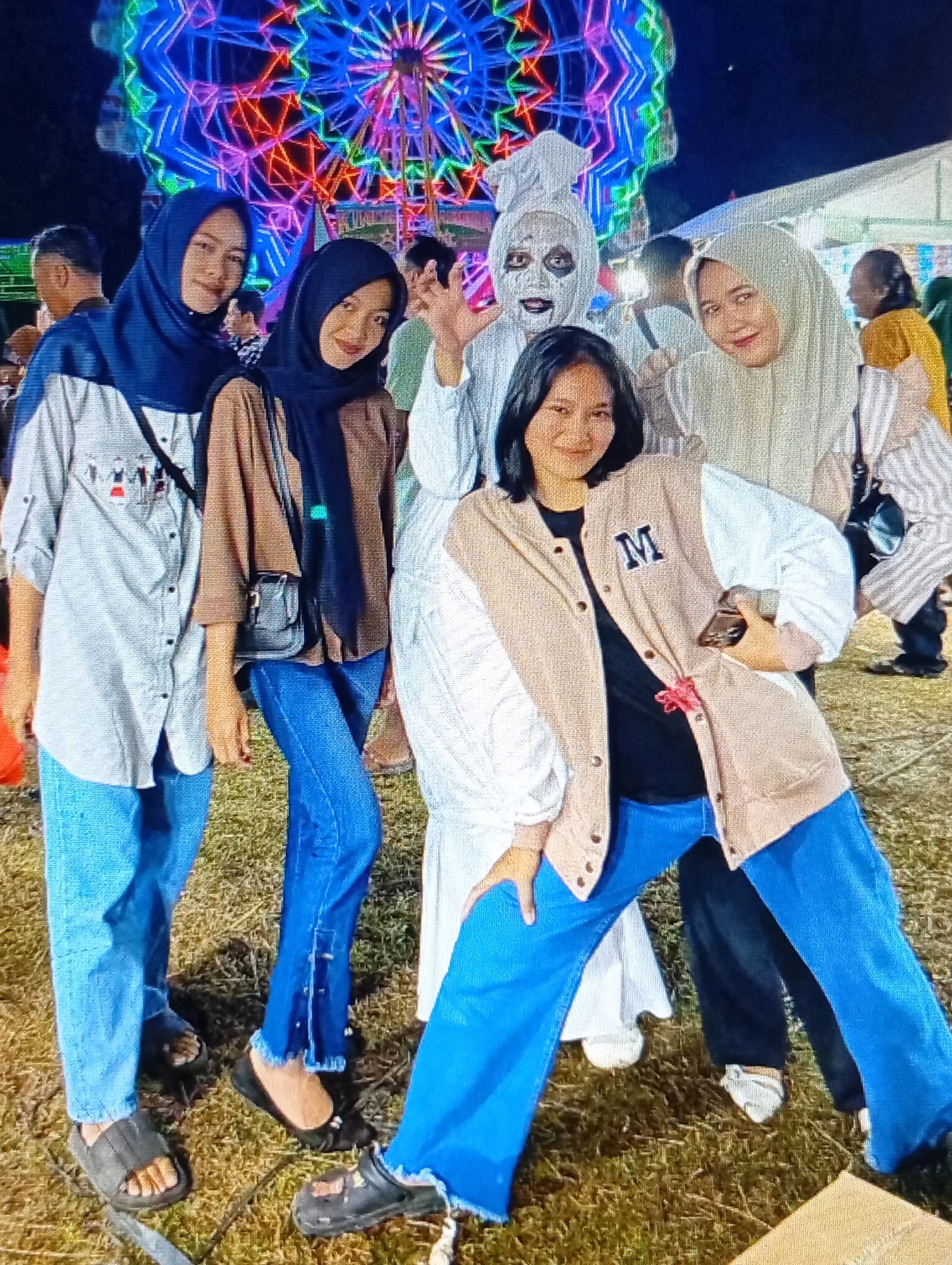This image captures a group of five people posing outdoors at night, with a brightly lit Ferris wheel visible in the background, likely at a fairground or carnival in Lhokseumawe, Indonesia. The central focus is on the group of four young women and one individual in a white costume with face paint.
From left to right:
The first woman is wearing a dark blue hijab, a light-colored button-up shirt with small red and black graphics on the chest, and light blue jeans. She is standing, facing the camera, with her left hand on her hip. She wears dark sandals.
The second woman is wearing a dark blue hijab and a brown long-sleeved shirt. She has a small black shoulder bag with a silver clasp. Her legs are visible, wearing blue jeans, and she is standing, facing the camera, with her left hand resting on her bag. She is wearing black flat shoes.
The third individual, positioned in the center, is dressed in a white ghost costume, complete with a white head covering and white face paint. This person is striking a playful pose, with one leg forward and hands outstretched.
The fourth woman is wearing a light-colored hijab and a light-colored jacket with white sleeves, a black t-shirt underneath, and the letter "M" embroidered on the left chest of the jacket. She is also striking a pose, leaning forward with her left hand holding a smartphone. She is wearing blue jeans and white shoes.
The fifth person, on the far right and partially obscured, appears to be a woman wearing a light-colored hijab and a striped top. She is standing and looking to her right.
The ground is covered in dry grass and some scattered leaves. In the background, the colorful lights of the Ferris wheel illuminate the night sky, creating a festive atmosphere. Tents are also visible in the background, suggesting a larger event. The lighting is primarily from the artificial illumination of the Ferris wheel and other nearby lights, casting shadows and highlighting the subjects. The overall scene suggests a recreational outing at a local festival.