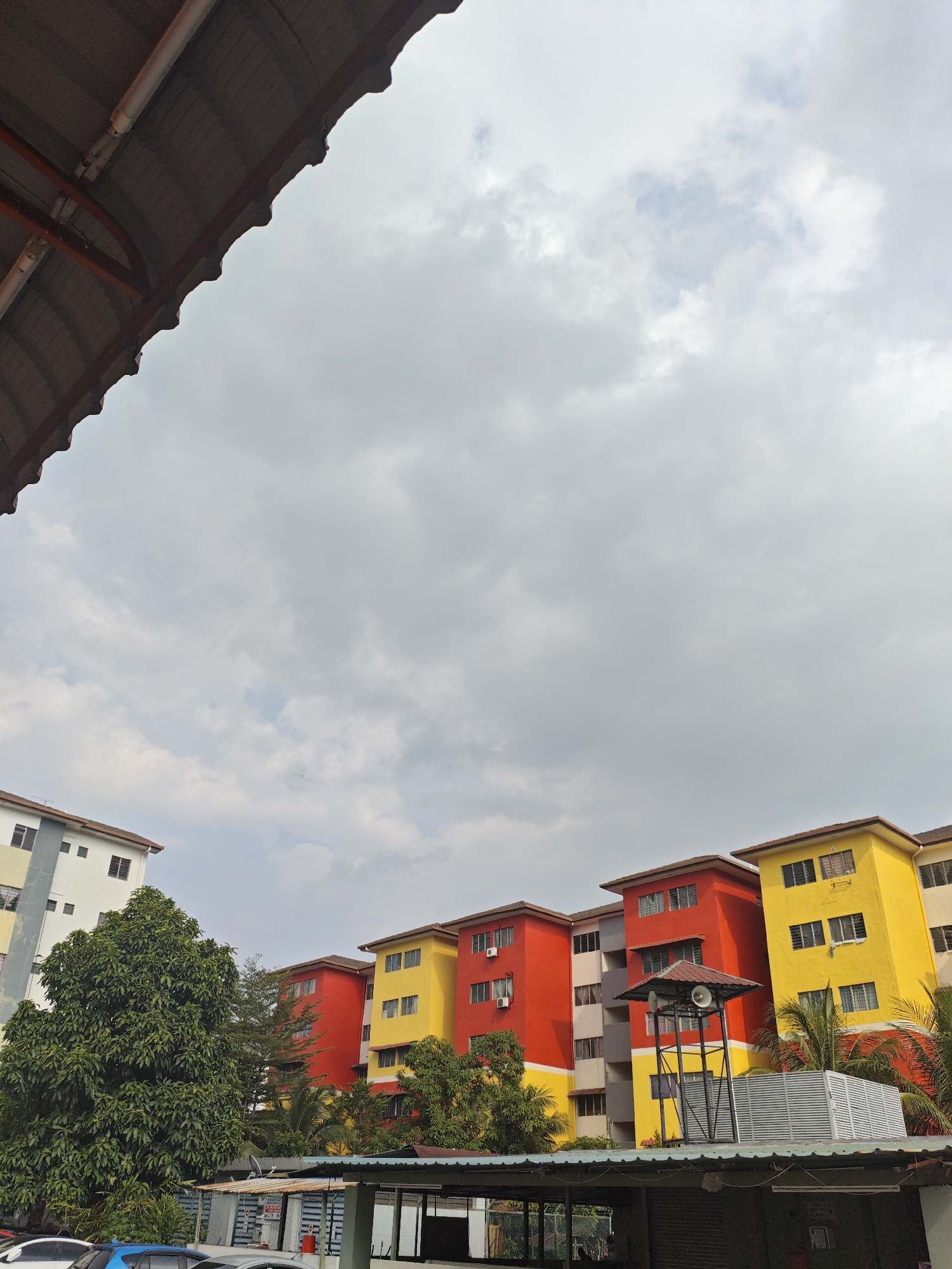 This is a photograph taken from a covered walkway looking out towards apartment buildings and trees under a cloudy sky. The walkway has a corrugated metal roof supported by orange beams. In the background, several apartment buildings are visible. The buildings are painted in bright colors, with sections of yellow, red, and off-white. Some windows and air conditioning units are visible on the facades. A large, leafy green tree is positioned to the left of the apartment buildings. A tall, dark metal structure with speakers is located in front of one of the buildings. The image appears to be taken in a residential area. The area is within the Kajang Municipal Council, Malaysia.