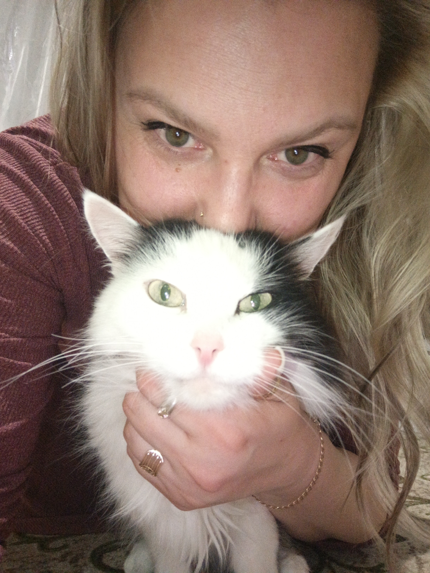 A close-up, intimate portrait captures a woman and her pet cat indoors, likely in Markovskiy, Russia. The blonde-haired woman, visible from her forehead to her upper chest, looks directly at the camera with warm, green eyes accentuated by dark eyeliner. She has a small gold nose stud and a minor facial piercing. She wears a dark red/maroon ribbed long-sleeved top. Her left hand gently cradles the cat's face, showcasing two gold-toned rings (one with a white stone) and a delicate gold chain bracelet on her wrist.
The domestic cat, a fluffy black and white breed, dominates the foreground. It features bright green eyes, long white whiskers, and prominent black markings around its ears and parts of its face. Its expression is calm and direct. The woman's face is positioned closely behind the cat's head, suggesting a strong bond and affection.
The background is simple, with a light-colored surface visible behind the woman's head and a patterned carpet or rug beneath the cat. The overall scene conveys a feeling of warmth and companionship. There is no visible text in the image.