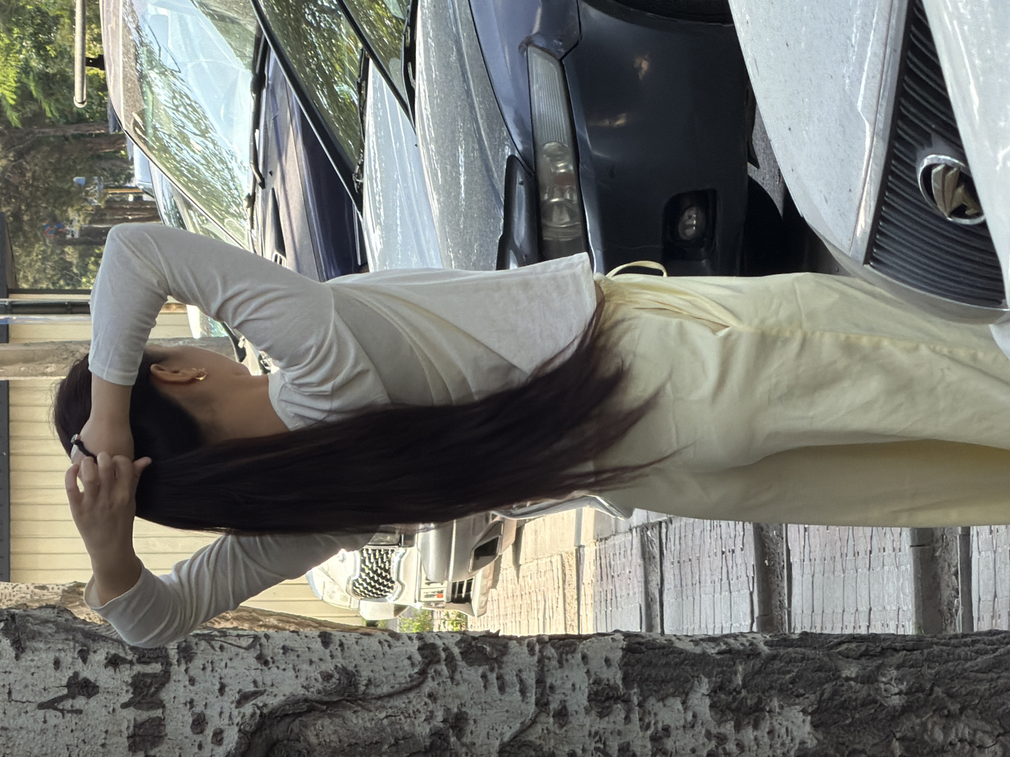 A woman with long dark hair pulled back in a ponytail, wearing a white long-sleeved shirt and light yellow pants, stands next to a tree trunk. She is seen from the back, with her arms raised and hands holding her hair. The scene appears to be outdoors, with parked cars visible in the background. The ground is paved with light-colored rectangular stones, and there is foliage and trees in the distance. The lighting suggests it is daytime.