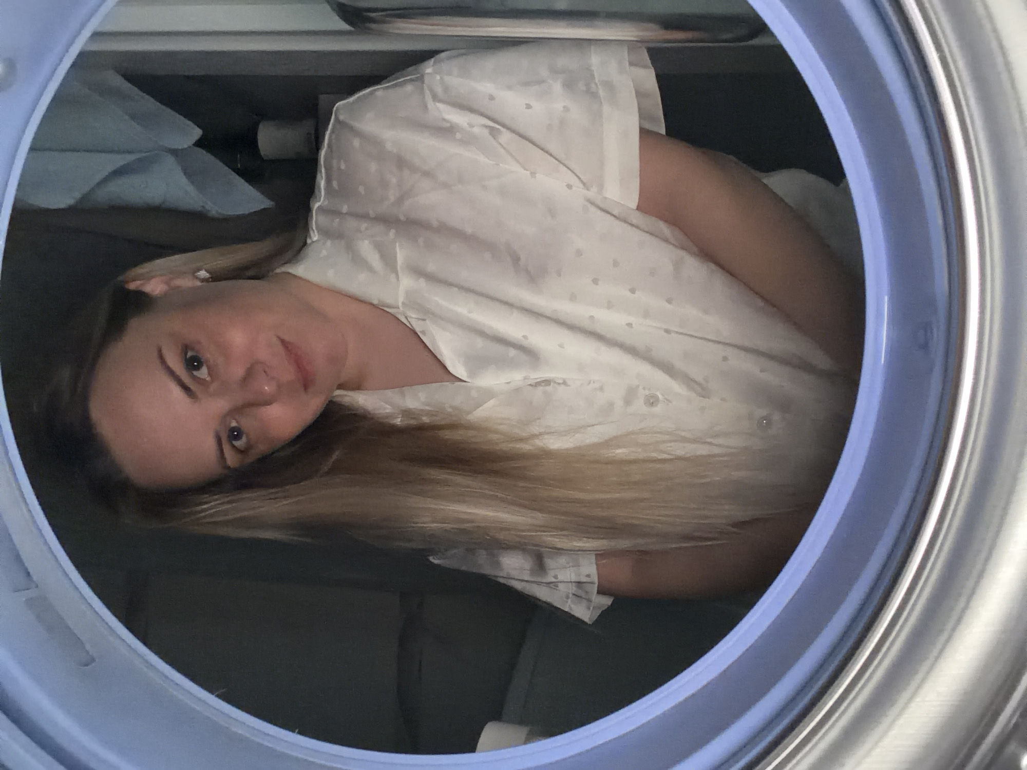 A woman with blonde hair looks toward the camera from inside a drum-shaped object with a blue rim. She is wearing a white, short-sleeved garment with small white polka dots. The interior of the drum is a dark grey or black. The upper portion of the background shows stacked light blue folded material. The image does not indicate that the scene is in Votkinsk, Russia.