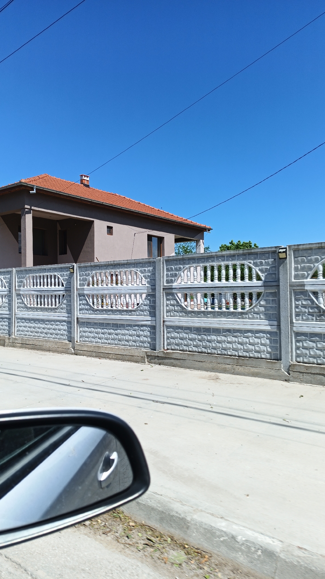 The image shows a residential scene in Boliartsi, Bulgaria. A light brown, two-story house with a red tile roof is partially visible behind a long, ornate gray concrete fence. The fence has decorative openings with vertical bars and oval shapes. Overhead electrical wires are strung against a clear blue sky. A side view mirror of a car is in the lower left corner, suggesting the picture was taken from a moving vehicle. The street in front of the fence is paved and clean. No people are visible. The scene is brightly lit, indicating a sunny day. Vegetation is sparsely visible in the background beyond the house.