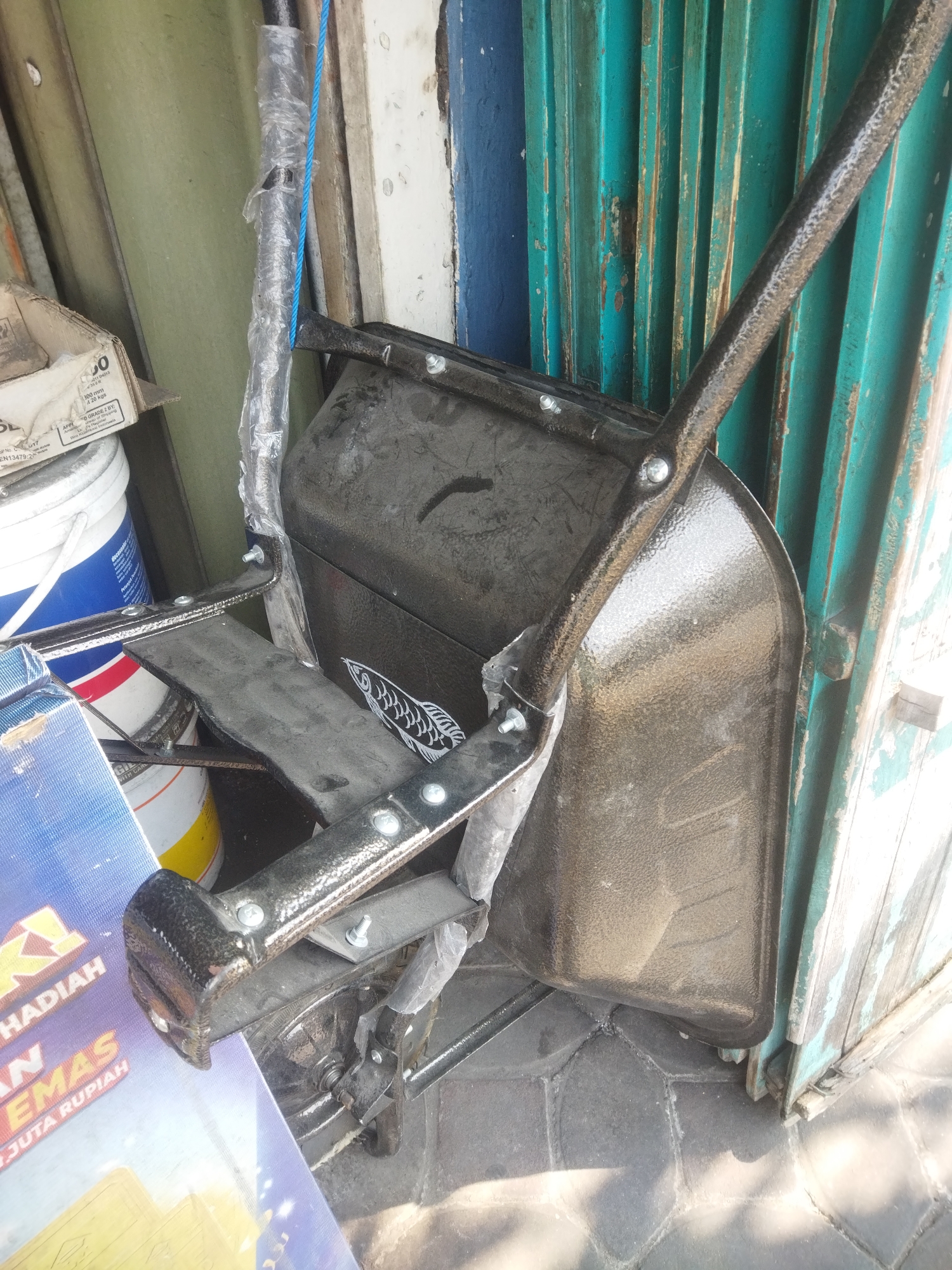 In Giripurwo, Indonesia, a black shovel with a textured metal scoop and a dark handle is leaning against a weathered teal-colored corrugated metal door. The shovel's handle is partially wrapped in clear plastic. To the left of the shovel, several large buckets are visible, one with a blue and white label. A cardboard box with printing, including the text "100 mm" and "20 KGS," is also present. A blue rope is tied to the shovel's handle and is taut. The ground is paved with hexagonal tiles, and some sunlight is casting shadows. The scene appears to be outdoors, possibly near a shop or storage area.