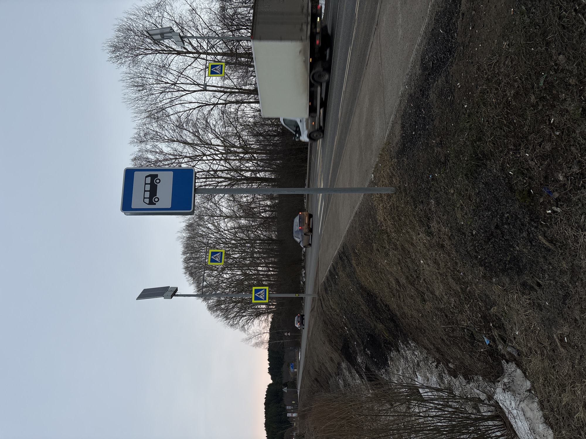 The image shows a roadside scene in Rusalevka, Russia. A bus stop sign is visible, along with pedestrian crossing signs featuring a yellow triangle with a black pedestrian symbol. A truck and passenger cars are present on the road. The environment includes trees without leaves, dry grass, and patches of snow on the ground. Solar panels are installed near the pedestrian crossing signs. The sky is overcast.