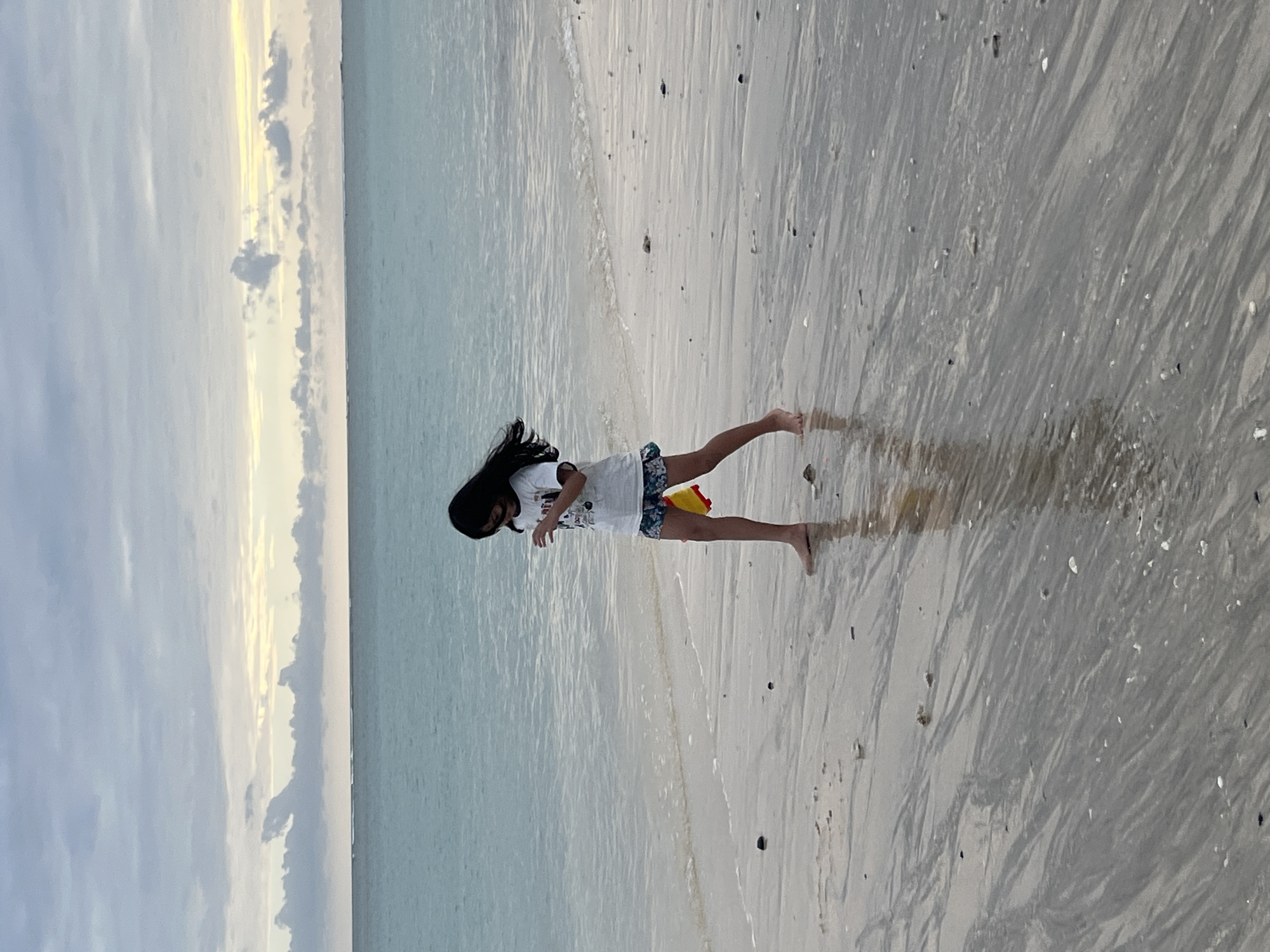 A young girl with long dark hair stands on a wet sandy beach, facing away from the camera. She is wearing a white t-shirt with a graphic print and patterned shorts. Her left leg is extended forward, and her right foot is slightly raised. A small red and yellow toy is held in her right hand. The sand is wet and reflects the sky, with small shells and dark specks scattered across it. The gentle waves of the ocean are visible to her left, and the water appears calm and a pale blue-green. The sky above is a mix of soft grey clouds and streaks of pale yellow light, suggesting either sunrise or sunset. The overall scene conveys a sense of peacefulness and a quiet moment at the beach. The location context is Kuta, Indonesia.