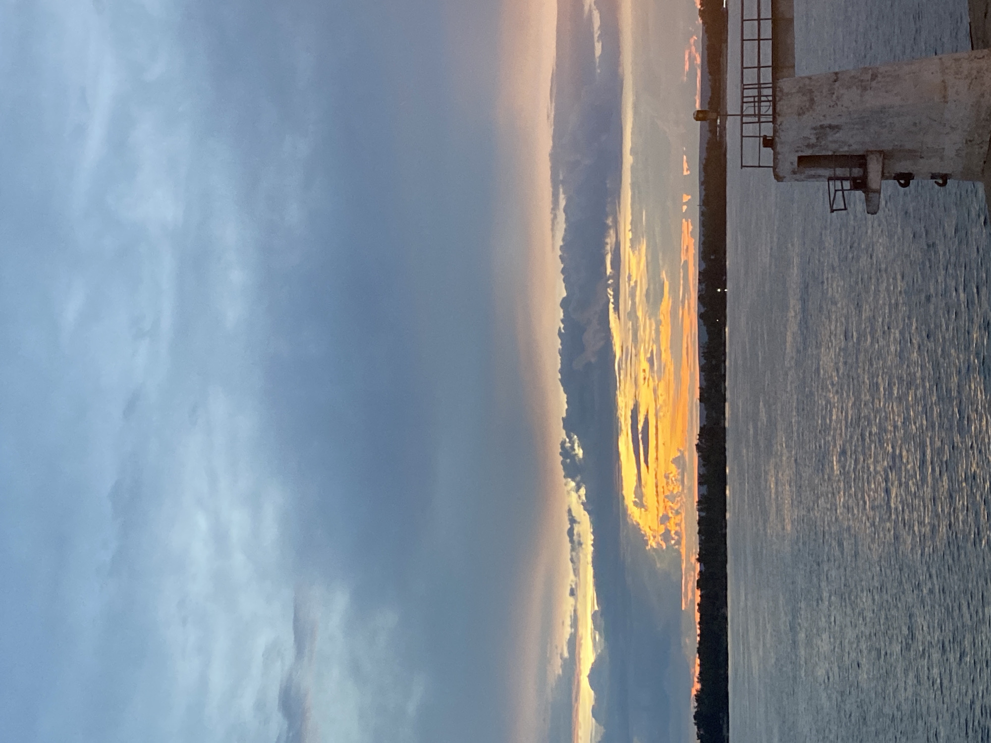 A photograph taken at dusk over a body of water, likely a river or bay, shows a picturesque sunset with colorful clouds. The sky transitions from pale blue at the top to vibrant hues of orange and yellow closer to the horizon, where a line of trees marks the distant shore. A concrete structure, possibly a pier or part of a bridge, occupies the lower right corner of the frame, adding a man-made element to the natural scenery. The water is calm, reflecting the sky's colors. The location is identified as Novokuybyshevsk, Russia. There are no people or animals visible in the image.