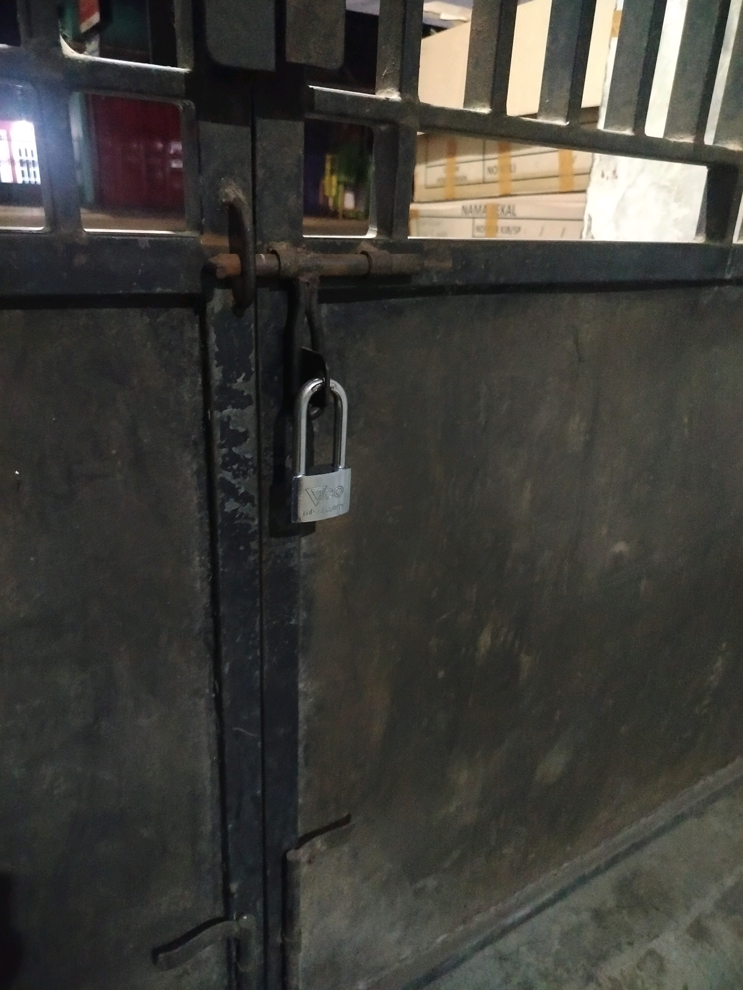 The image shows a close-up view of a black metal gate secured with a silver padlock. The gate appears to be closed and is constructed from vertical bars and solid panels. A latch mechanism is visible, through which the padlock is attached. In the background, through the bars of the gate, stacks of cardboard boxes can be seen. The scene is dimly lit, suggesting it might be evening or night. There is no indication of the city of Kroya, Indonesia in the image.