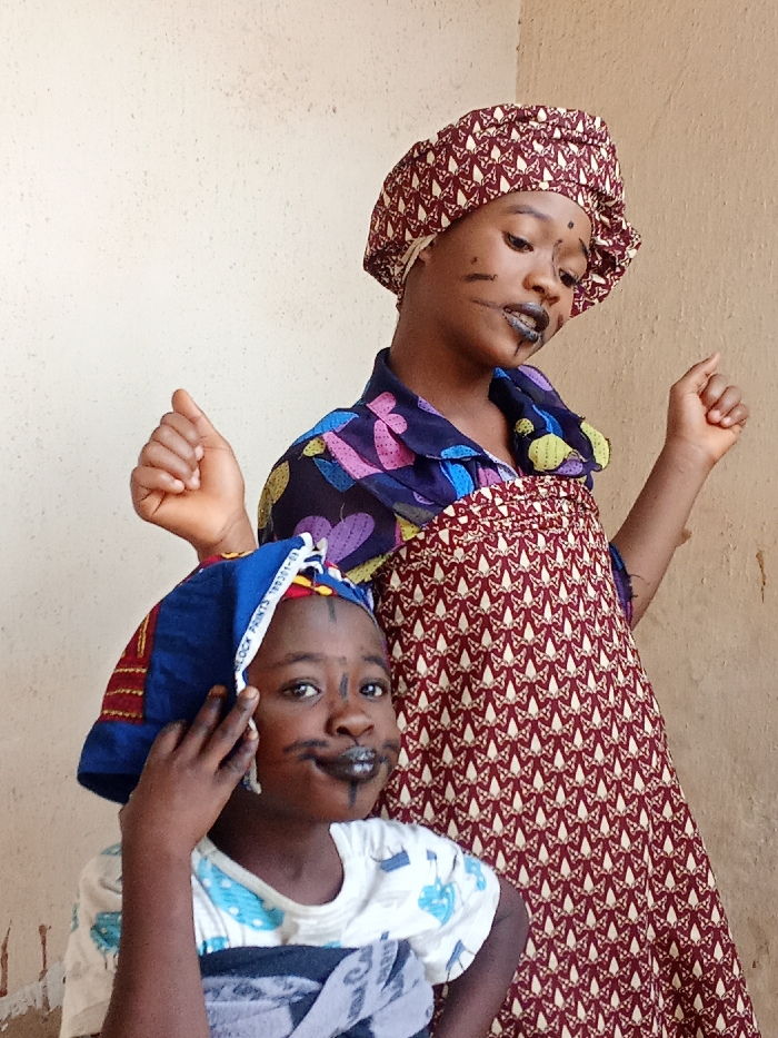 Two young girls pose indoors against a beige wall. The girl in the front wears a blue head covering, a white shirt with blue patterns, and has dark lines drawn on her face resembling a mustache and eyebrows. The girl behind her has a patterned burgundy and white dress and a matching head covering. She also sports similar facial markings. Both girls are raising their hands, appearing playful and lively.