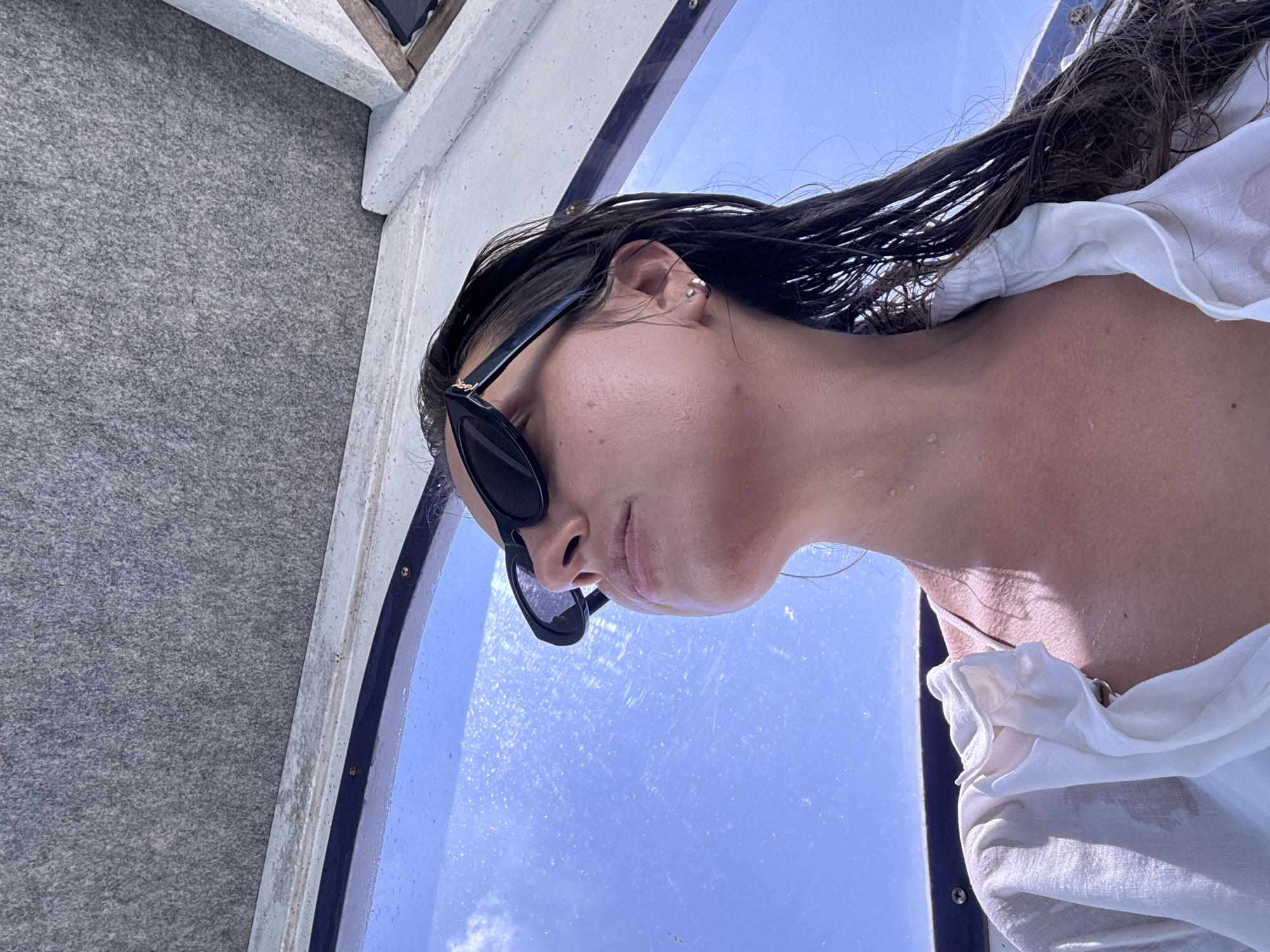 A woman with wet, dark hair and sunglasses is seen in an upward-facing shot with a view of the sky through a blue-tinted window. She is wearing a white, loose-fitting top. The background includes a section of a gray wall or surface on the left and a white structure above. The image appears to be taken in Unknown, Indonesia.