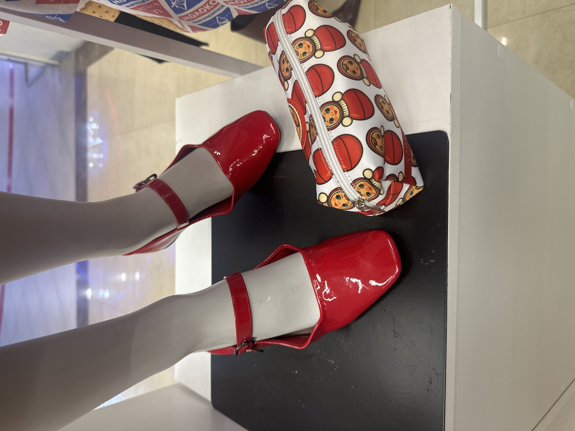 The image shows a display with two red, patent leather Mary Jane shoes on what appears to be mannequin legs. The shoes are styled with a strap across the instep. A cylindrical pencil case adorned with a pattern of Russian nesting dolls rests on a black and white surface next to the shoes. The display is set in a retail environment, with a window behind it reflecting the outside world. The location in the city of Yerevan, Armenia is not possible to identify from the information present in this image.