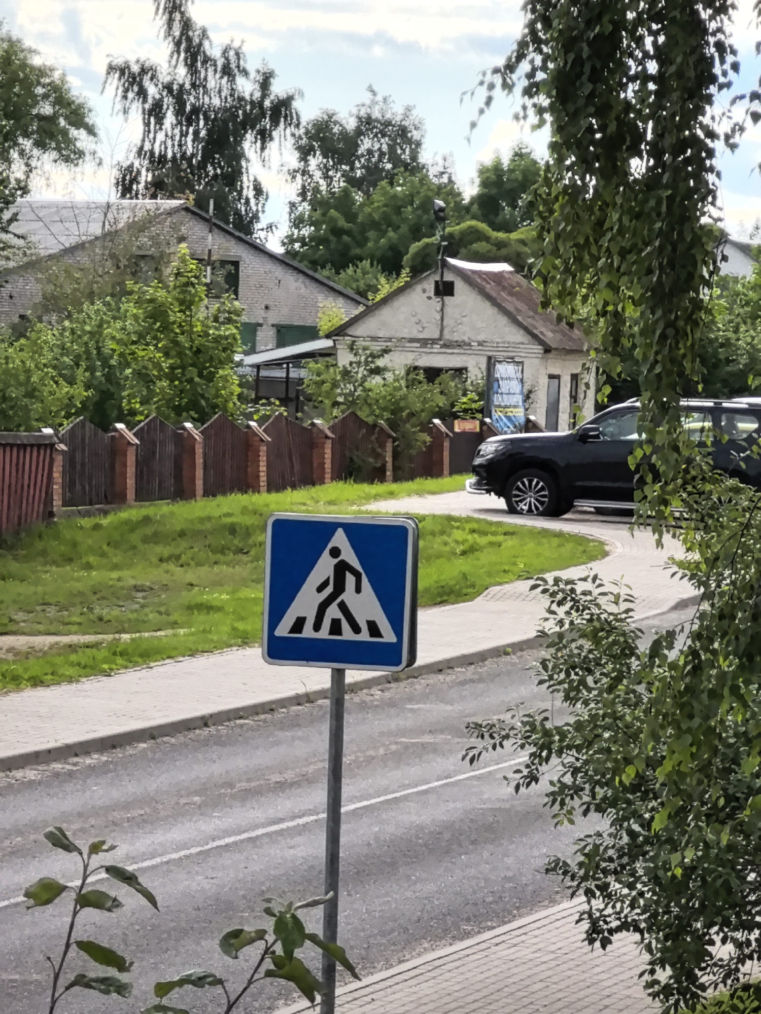 In an outdoor, daylight scene, a black SUV is parked on a paved street near a crosswalk sign. The crosswalk sign, a blue square with a white triangle inside depicting a pedestrian figure, is positioned on a metal pole in the foreground, partially obscuring a tree. The street curves to the right, and the SUV is situated on this curve. A sidewalk with a brick pattern runs alongside the street. Behind the sidewalk, a grassy area leads to a wooden fence with brick posts. Further in the background, two buildings are visible, one appearing to be a house with a gray roof and white brick walls, and the other a smaller structure with a weathered roof. Lush green trees and foliage surround the buildings and fence. The sky above is partly cloudy.