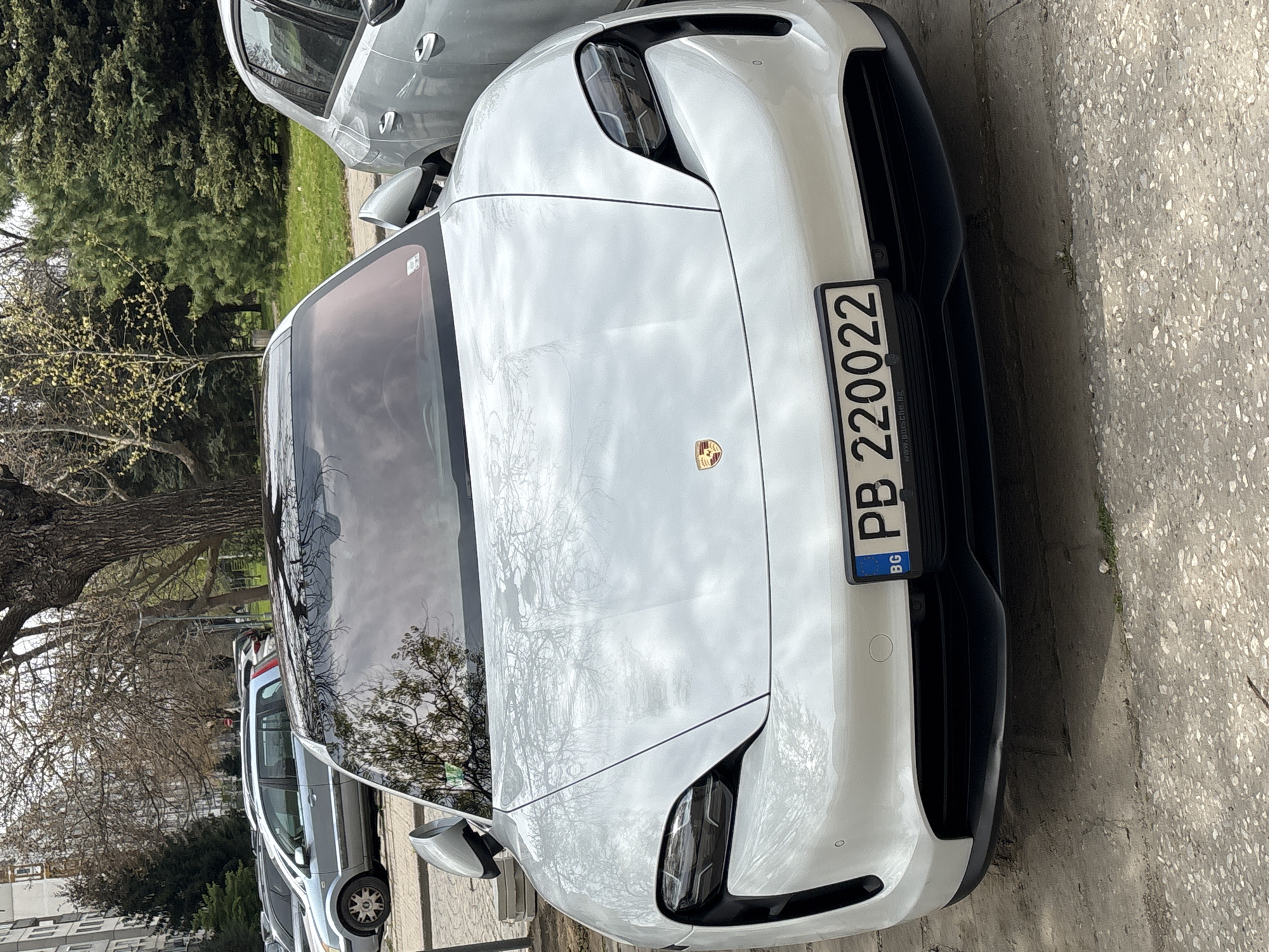A white Porsche Taycan is parked on a street in Plovdiv, Bulgaria. The car has a Bulgarian license plate, PB 220022. The car is parked near a sidewalk with trees and other cars visible in the background.