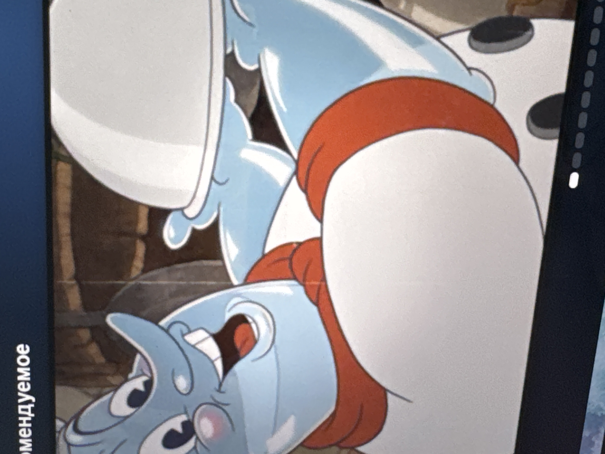 The image is a close-up of an animated character with blue skin, a long blue nose, and rosy cheeks. The character is wearing a red scarf around its neck and a white outfit with black polka dots. It is holding a white domed lid, suggesting it is serving food or presenting something. The background appears to be an indoor setting, possibly a kitchen or dining area, with wooden textures. There is text in the top left corner that reads "рекомендуемое" which is Russian for "recommended". There is no visible indication of the city of Riga, Latvia, in the image.