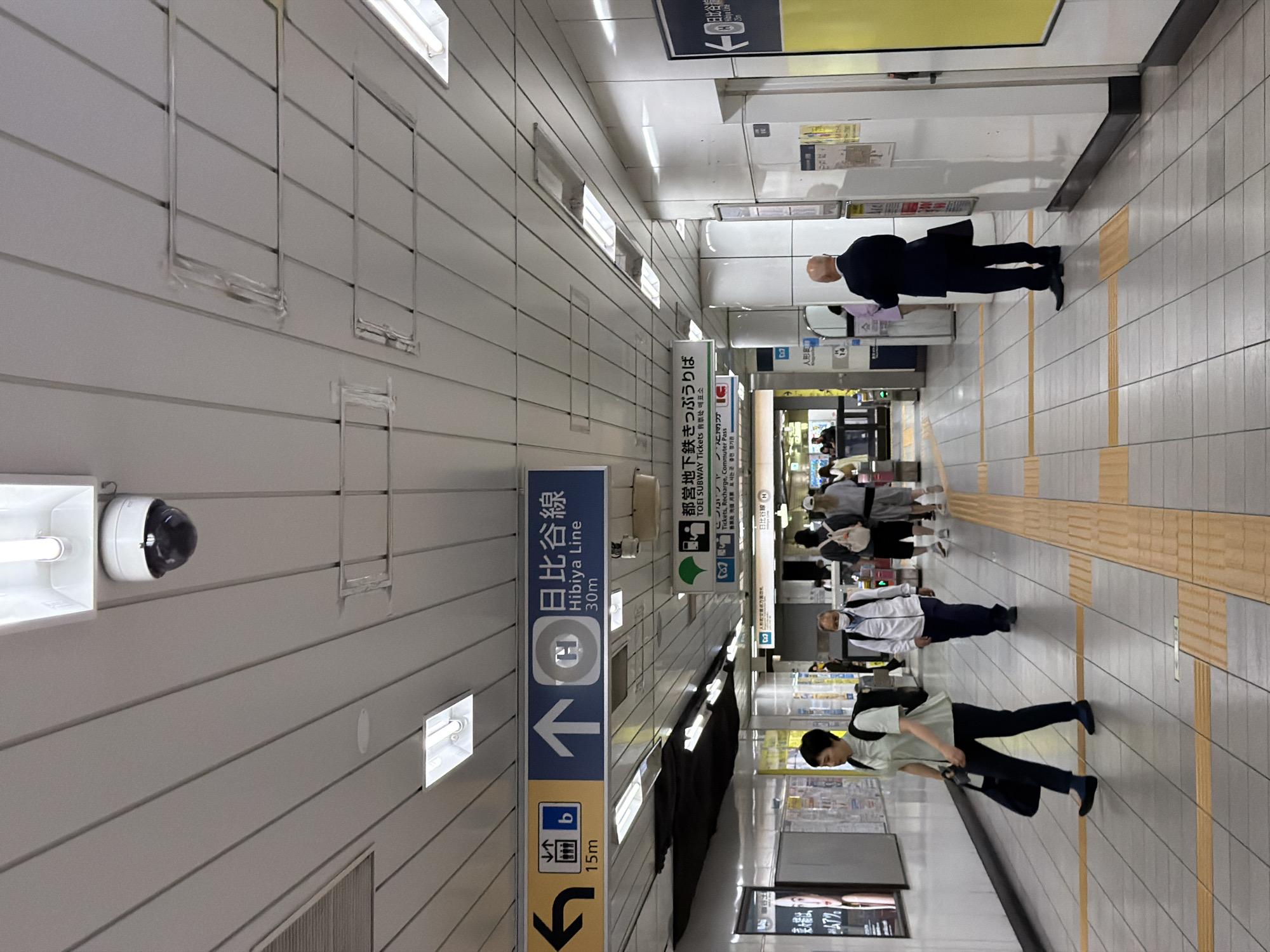 The image shows a subway station interior in Chuo, Japan. The scene is a long, brightly lit corridor with a high, slatted ceiling. Fluorescent lights are embedded in the ceiling, and a security camera is visible near the center.
Several signs are suspended from the ceiling, providing directions in Japanese and English. One prominent sign indicates "Hibiya Line 30m" with an upward arrow. Another sign points left with an arrow and displays "15m". A green sign with a stylized tree and text including "TOEI SUBWAY Tickets, Rechange, Commuter Pass" is also visible.
Several people are walking through the station. In the foreground, a woman with a black backpack walks to the left, carrying a dark bag. Further down the corridor, a man in a white jacket and dark pants walks towards the viewer, wearing a face mask. Another man in a dark suit stands facing away from the viewer, holding a black briefcase. Other individuals are seen in the background, some walking and others standing.
The floor is tiled with a light gray and white checkered pattern, with yellow tactile paving strips indicating the edge of the platform. The walls of the station are white and appear to be made of panels. Advertising posters and information displays are mounted on the walls. The overall atmosphere is that of a busy, functional public transportation hub.