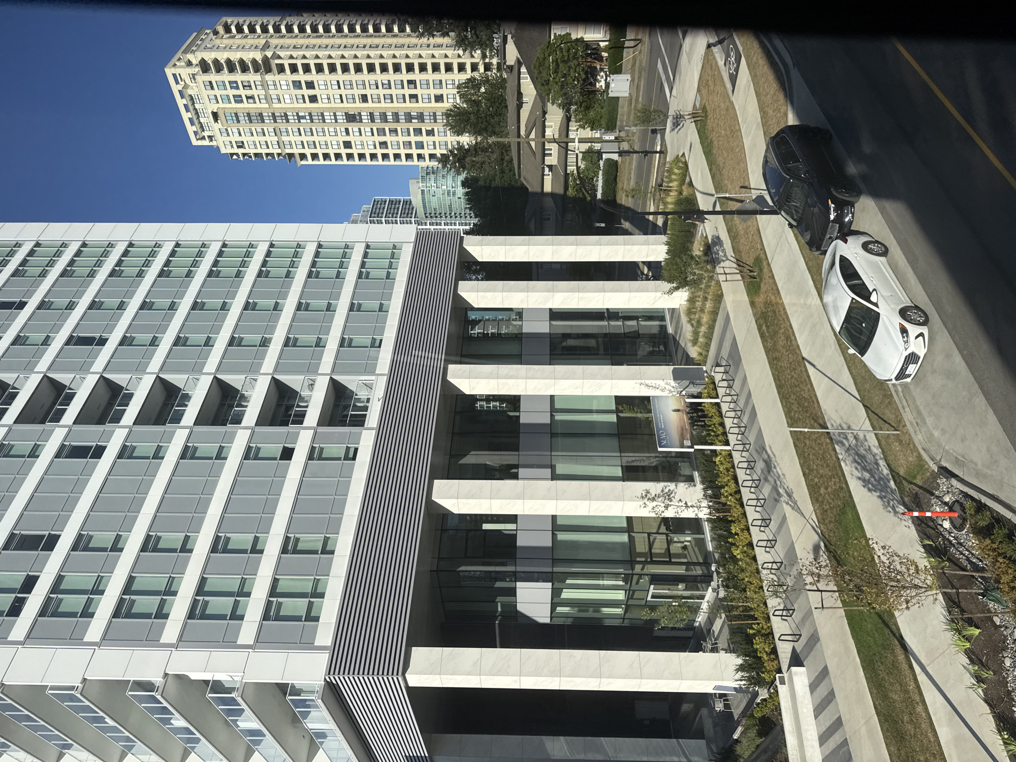 This image captures an urban scene in Burnaby, Canada, featuring a contemporary apartment building with a glass and white facade, characterized by multiple balconies and rows of windows. The building's base has a commercial or lobby area with large glass windows and prominent white columns. Adjacent to this building, a taller, light-colored high-rise building stands, also featuring numerous windows and balconies. In the foreground, a paved sidewalk runs alongside the buildings, bordered by greenery and a bicycle rack. Two cars, a white sedan and a black SUV, are parked on the street. The environment appears to be a sunny day with a clear blue sky. The overall scene depicts a modern residential or mixed-use development area within a city.