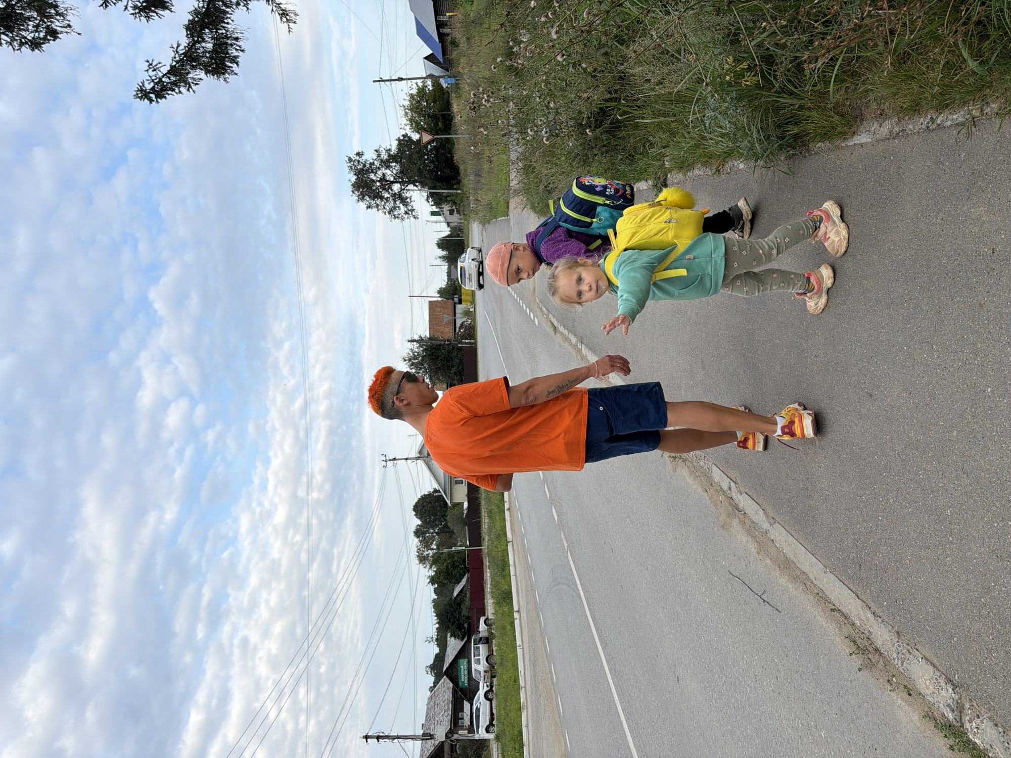 In Chaikovsky, Russia, on a paved road with a grassy shoulder and scattered weeds, a young man with bright orange hair walks away from the camera on the right side of the frame. He is wearing an orange t-shirt and navy blue shorts. To his left, on the sidewalk, a young girl with blonde hair and a mint green hoodie, carrying a yellow backpack, extends her left hand towards the viewer as if to stop or greet. Behind her, a slightly older child wearing a pink hat and a purple jacket with a blue backpack turns their head to look at the viewer. The background reveals a rural or suburban setting with wooden houses, trees, and power lines. Two white cars are parked on the left side of the road. The sky is overcast with scattered clouds.