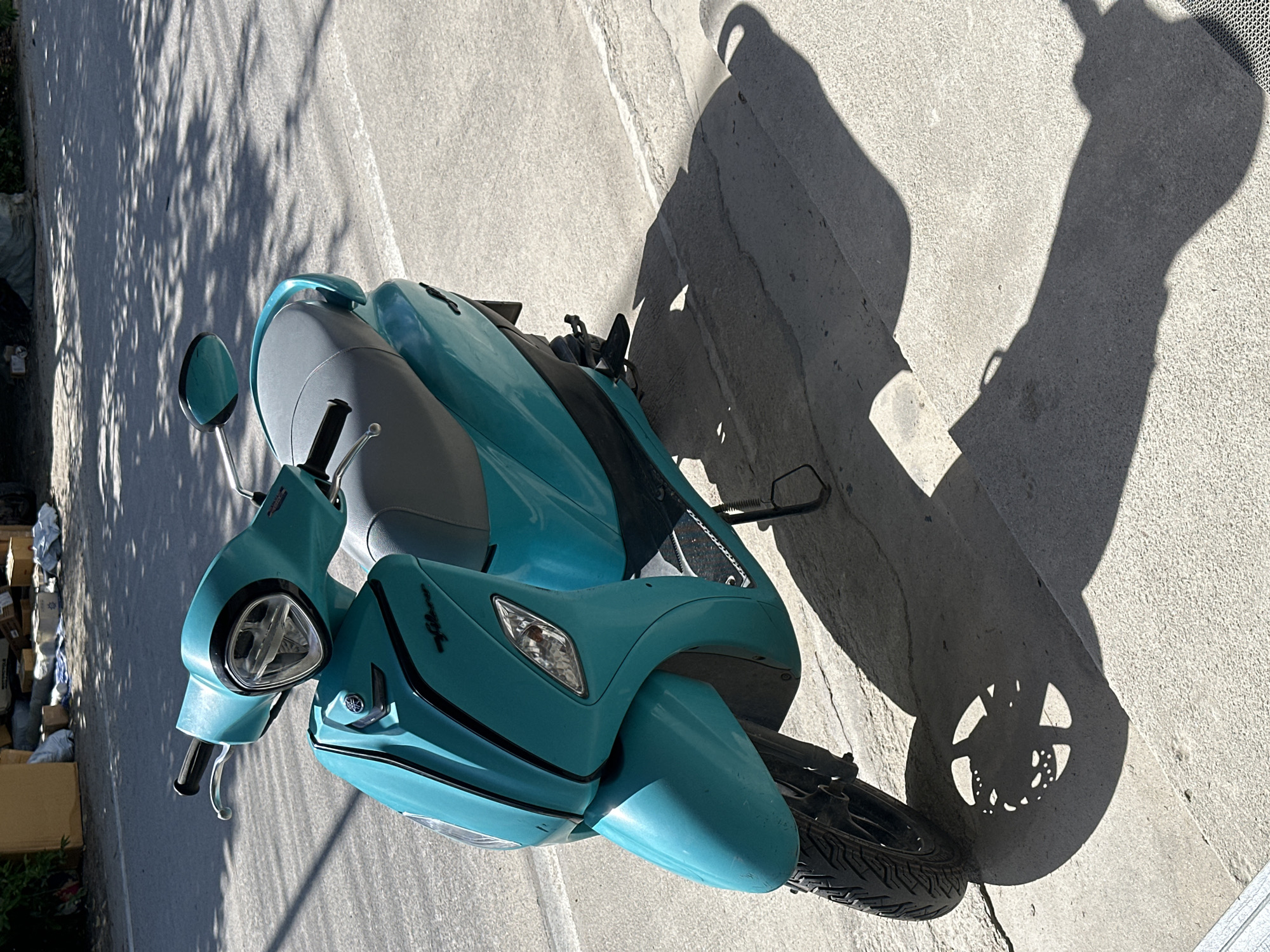 A teal-colored scooter is parked on a concrete sidewalk next to a road, casting a long shadow. The scooter has a gray seat, a prominent headlight, and a Yamaha logo on its front. The background shows a road with dappled sunlight filtering through trees, and some boxes and bags are visible on the side of the road, possibly indicating a commercial or storage area. The scene appears to be in a public outdoor space in ตำบลเกาะพะงัน, Thailand.