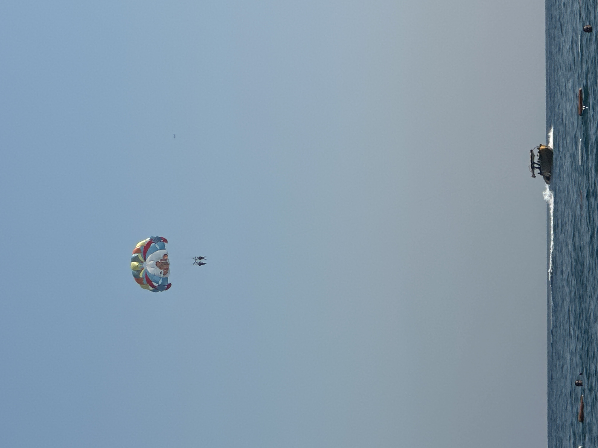 In the city of Boreti, Montenegro, two individuals are parasailing high above the clear blue sea. A colorful parasail, a vibrant mix of red, yellow, blue, and white, billows in the sky, supporting the two figures suspended beneath it. Below them, a motorboat navigates the water, leaving a white wake. The sea is a deep blue, with gentle waves. In the distance, the horizon is a pale line against the clear sky, with a tiny airplane visible as a speck. On the water's surface, there are a few buoys and a person visible near one of them. The overall scene depicts a recreational activity taking place on a sunny day.