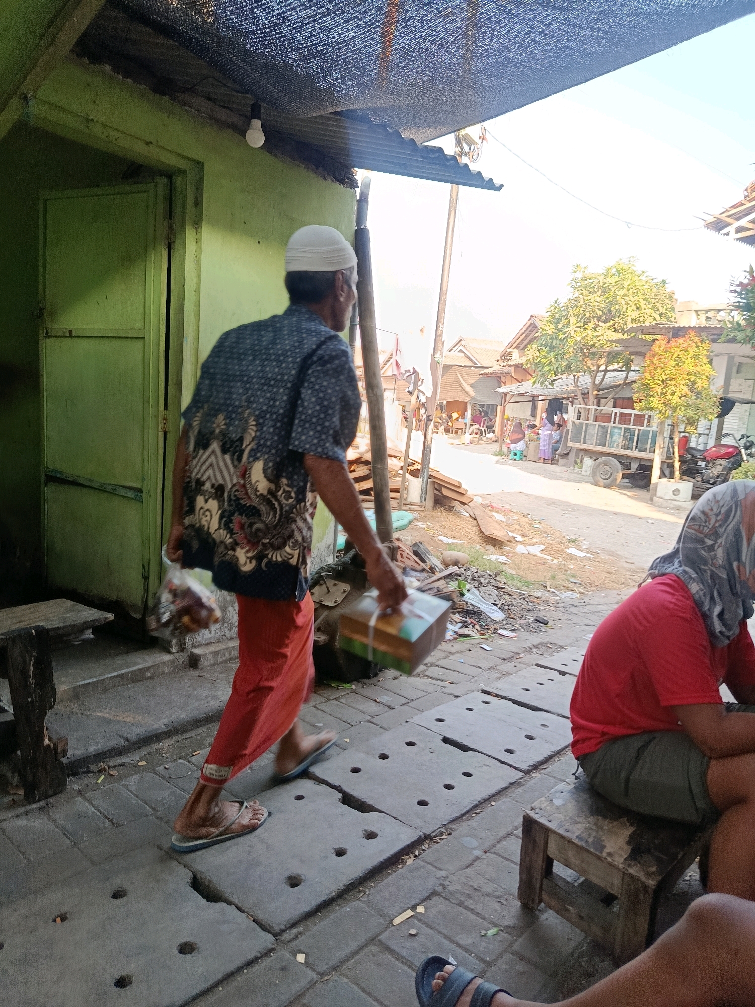 In the city of Jobokuto, Indonesia, a man wearing a white cap, a dark patterned shirt, and orange pants walks on a paved walkway carrying a box and a plastic bag with items inside. To his right, a person in a red shirt and gray head covering sits on a wooden stool. The background reveals a street with buildings, a truck, a motorcycle, and trees under a bright sky. A green building with an open door is visible on the left side of the image.