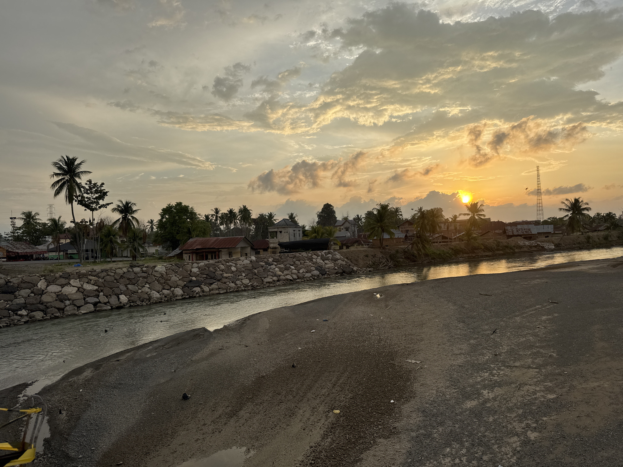 The image depicts a sunset scene in Meunasah Pupu, Indonesia. A wide river flows from right to left across the frame. The riverbanks are a mix of natural earth and a constructed stone wall on the left bank. On the left bank, several buildings, including houses with reddish-brown roofs and some with concrete structures, are visible behind the stone wall. Palm trees are scattered throughout the landscape.
The sky is filled with dramatic clouds, illuminated by the setting sun on the right side of the image. The sun, a bright orb, is low on the horizon, casting a warm, golden light across the sky and reflecting on the water. A tall, slender radio tower stands against the sunset on the right. The foreground consists of a sandy and pebbly area, with some scattered debris. The overall atmosphere is tranquil, with the natural beauty of the sunset contrasting with the human settlement along the river.
