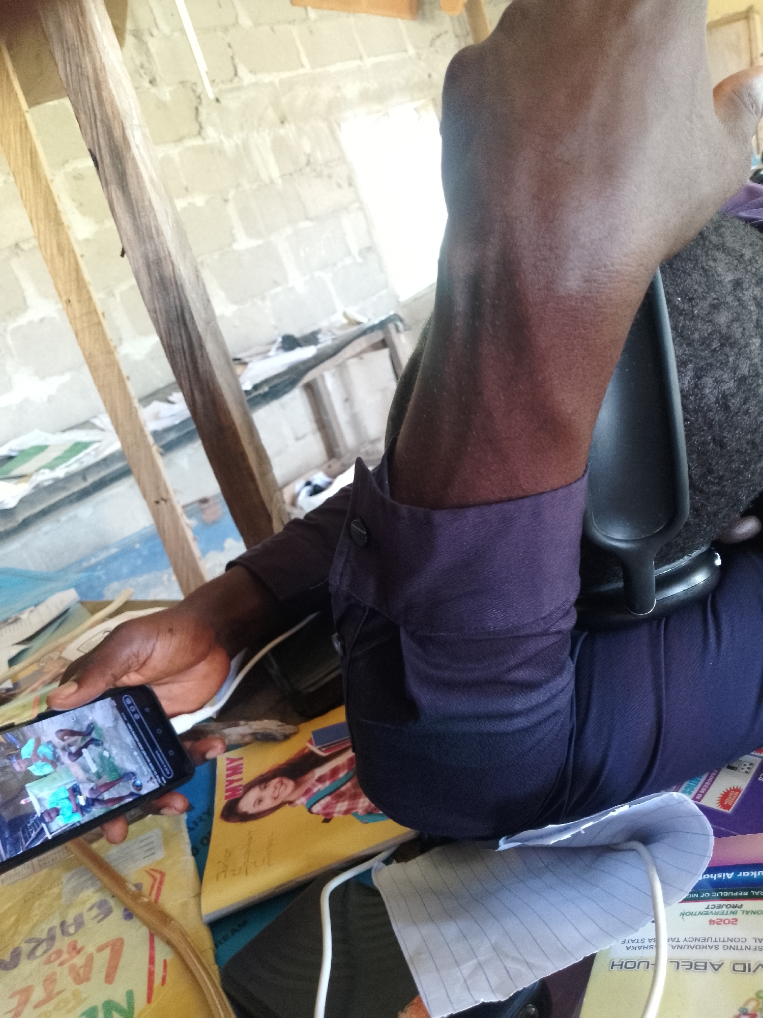 A person is sitting down, holding a smartphone in their left hand and looking at the screen. The person is wearing a dark purple long-sleeved shirt and dark blue pants. A black office chair is visible behind them. The background shows a wall made of concrete blocks, some wooden beams, and papers or books scattered around. A phone charger cable is plugged into the phone. The phone screen displays a video or image of several people in a green outdoor setting.
The scene appears to be indoors, possibly a workshop or office space, with some construction elements visible. The lighting suggests it is daytime. There are no clear indicators of the weather or specific emotions.
Visible text on the papers and books includes: "MYMY", "2024", "PROGRESS IN", "NATIONAL INTERVENTION PROJECT", "ENSING SARDAUUA", "STATE", "DAVID ABEL - UOH", "SHAKA". There is also text on the phone screen, but it is too blurry to read. The general context suggests it might be in Serti, Nigeria.