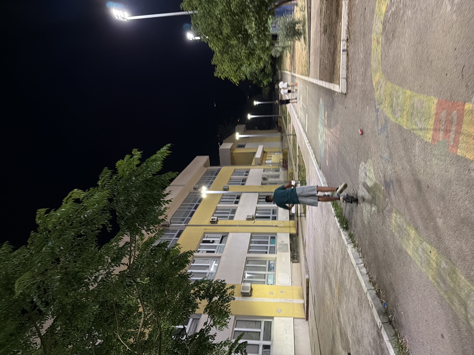 In the city of Budva, Montenegro, at nighttime, a young person rides an electric scooter on a paved walkway in front of a yellow and white building with multiple windows. The walkway is adorned with colorful chalk drawings, including a yellow and red arch. Several other individuals, including children, are visible further down the walkway, some appearing to be walking. The scene is illuminated by streetlights. Lush green trees with dense foliage border the walkway and the building. The sky is dark, indicating it is night.
