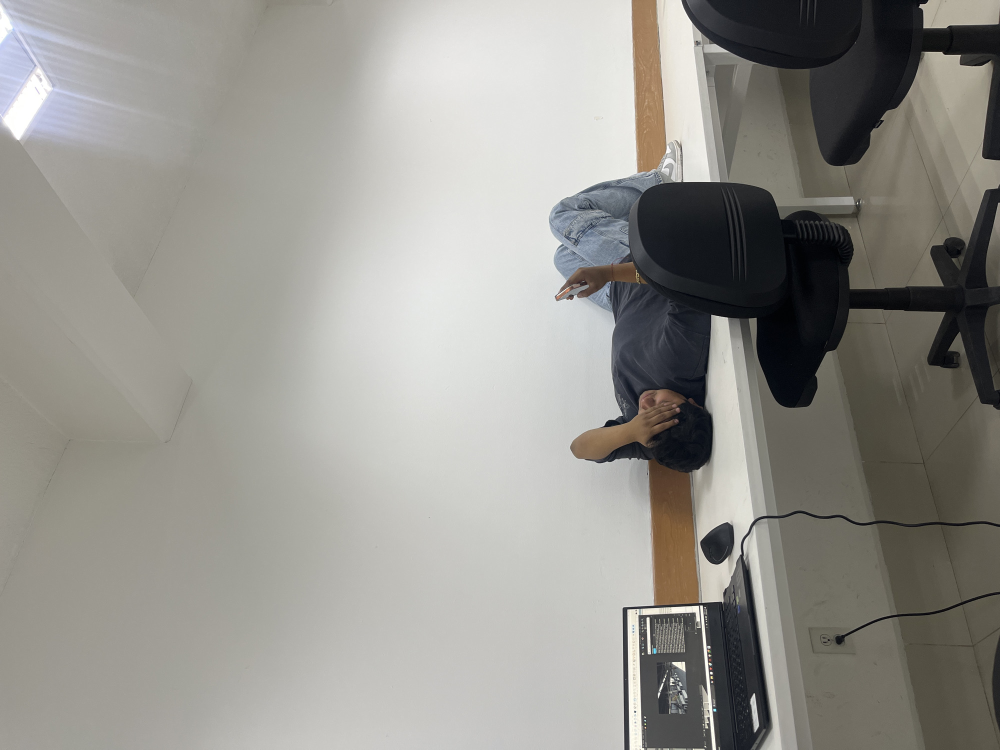 A person is lying on the floor of a room in San Andrés Cholula, Mexico, next to a desk with a laptop and computer mouse on it. The person is wearing a grey shirt, light blue jeans, and white and grey shoes. The person is holding a white object. A black office chair is nearby. The room has white walls and a white ceiling with a window.