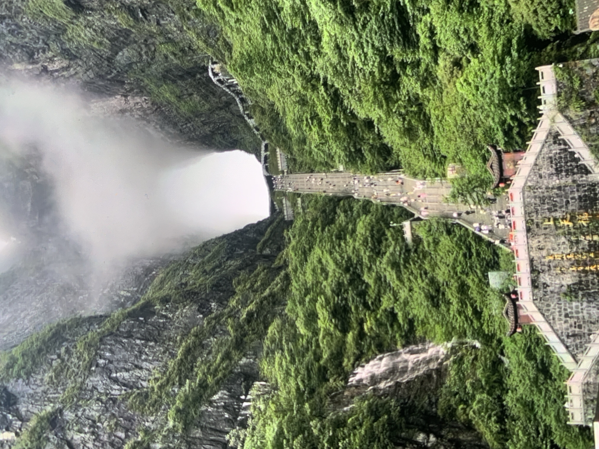 This image depicts a vast stone staircase ascending a steep, lush green mountainside. The staircase is densely populated with numerous people walking upwards. The mountains are heavily forested with vibrant green trees, and on the left side, a waterfall cascades down the rocky terrain. At the base of the staircase, a traditional Chinese-style pavilion with red roofs and white railings is visible, adding an architectural element to the natural landscape. The scene is partially obscured by mist and clouds at the top, creating a dramatic and awe-inspiring atmosphere. The image is not from Stavropol, Russia, but rather appears to be a natural wonder or historical site, possibly in Asia, given the architectural style.