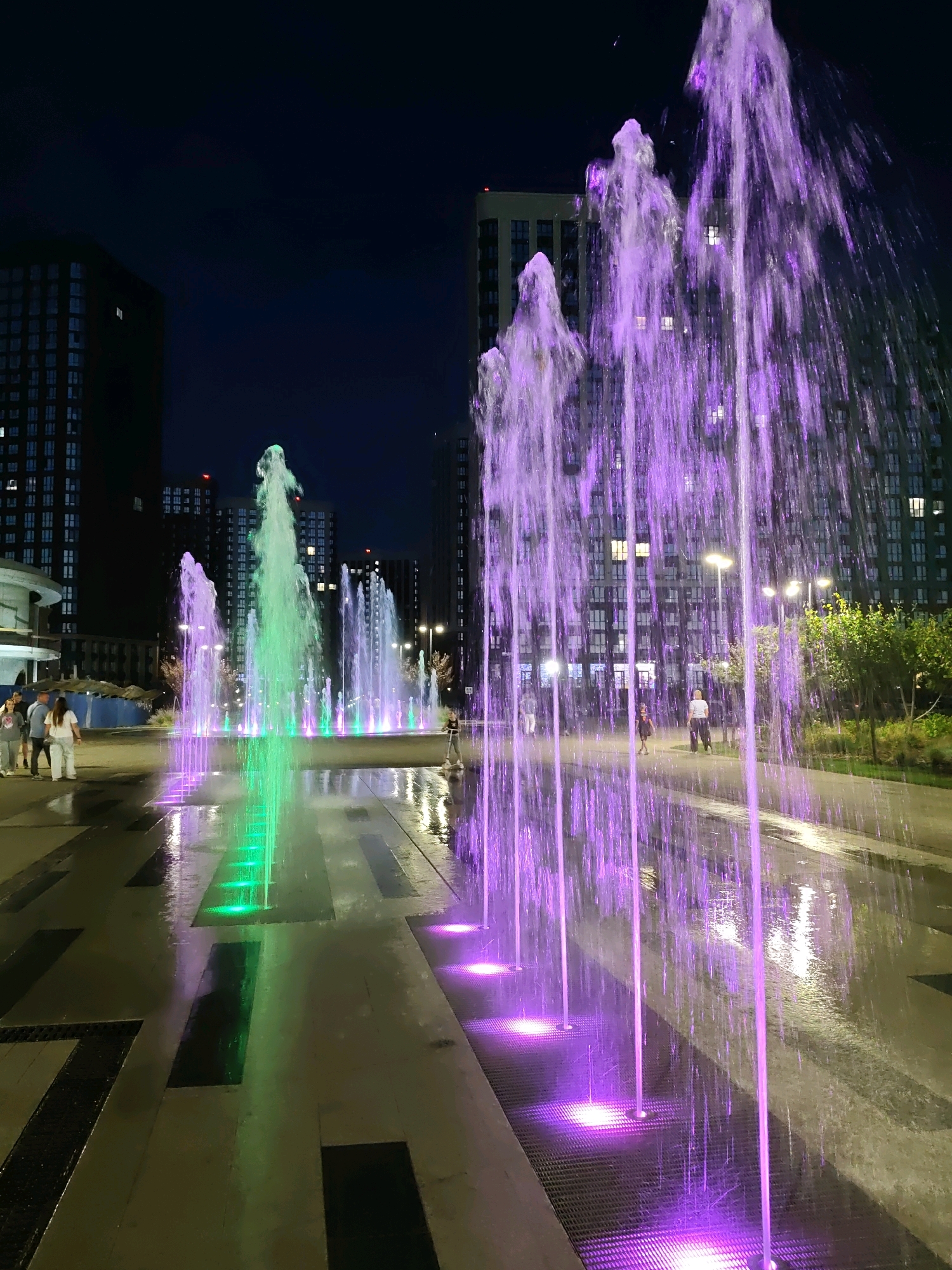 This is a nighttime photograph of a modern urban plaza featuring a light-up fountain in Rostov-on-Don, Russia. The fountain's water jets are illuminated in vibrant green and purple hues, creating a striking visual effect against the dark sky and the surrounding contemporary buildings. The wet pavement reflects the colorful lights, adding to the dynamic atmosphere. Several people are visible in the scene: a group of individuals stands on the left side of the plaza, while a person on a scooter is moving across the right side. The architecture suggests a newly developed residential or commercial area, characterized by tall, multi-story buildings with illuminated windows. The overall impression is one of a lively and aesthetically pleasing public space at night.