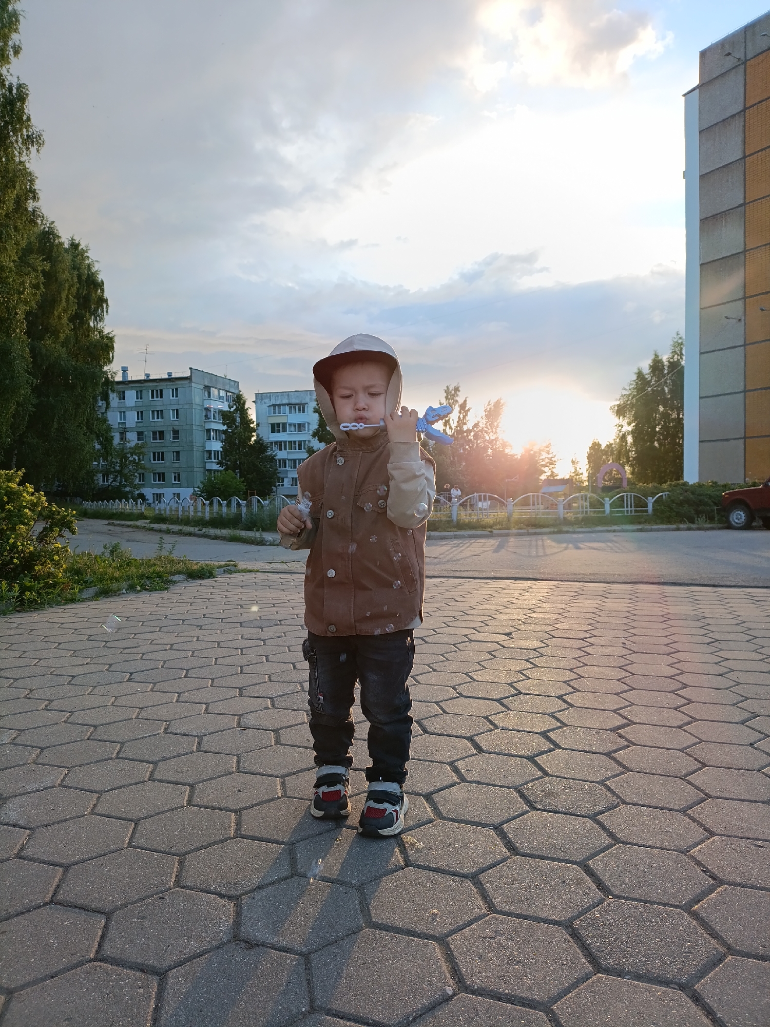 A young boy is standing on a paved area made of hexagonal tiles, holding a blue bubble wand to his mouth. He is wearing a hooded brown vest over a light-colored long-sleeved shirt and dark pants. The boy's expression is focused on blowing bubbles.
In the background, there are apartment buildings and trees. The sun is setting, casting a warm glow and long shadows across the scene. The sky is partly cloudy, with a bright sunset visible. The setting appears to be a residential area, possibly a courtyard or park, in Chaikovsky, Russia, given the architectural style of the buildings. The overall mood is peaceful and captures a moment of simple childhood joy. There is no visible text in the image.