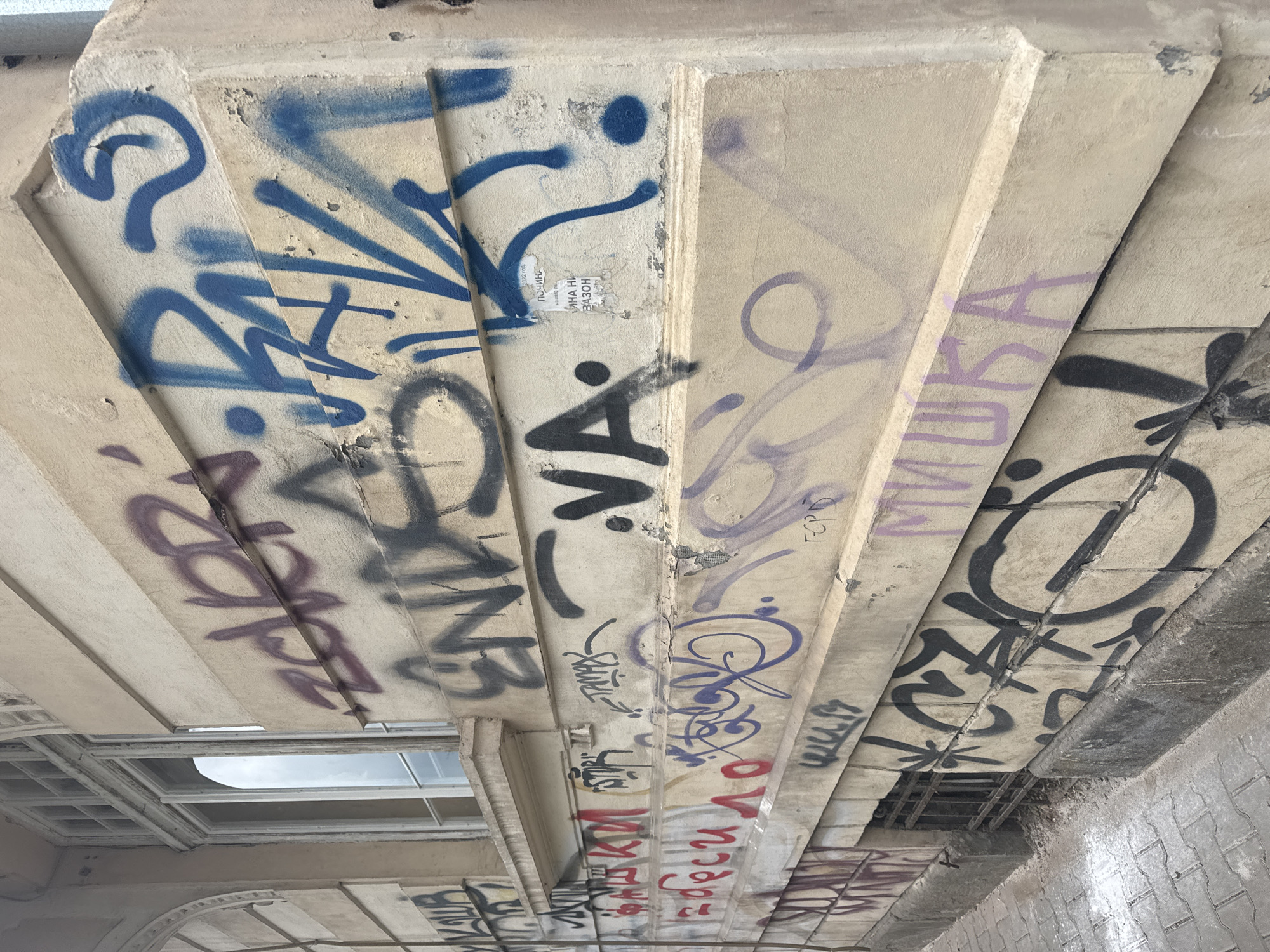 The image shows the underside of a concrete structure, possibly a bridge or overpass, heavily covered in graffiti. The graffiti consists of various tags, words, and abstract designs in multiple colors, including blue, purple, black, and red. The structure is made of segmented concrete beams or panels.
The presence of text in Cyrillic script, as well as the phrase "Миша Ни вазон" suggests the location is Sofia, Bulgaria.
There is also a partially visible window or opening within the structure, revealing the sky. The surrounding architecture is made up of concrete. There are also pieces of metal framing in the lower right corner.