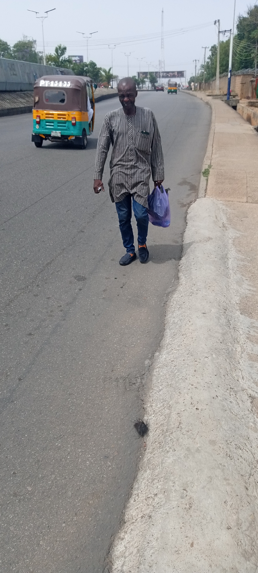 The image shows a man walking on a road in Jos, Nigeria. He is wearing a long-sleeved, striped shirt, jeans, and black shoes. He is holding a purple plastic bag in his right hand and a phone in his left. Behind him, a "Keke" (tricycle taxi) is driving on the road. In the background, there are buildings, trees, and overhead power lines. There is also what appears to be road debris on the road.