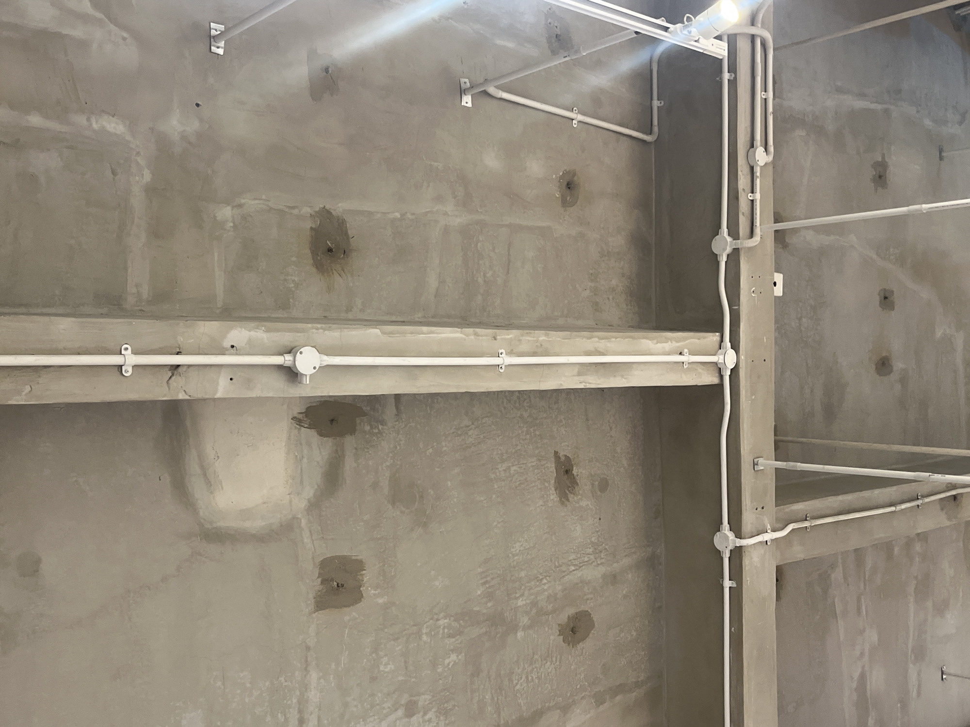 The image depicts an unfinished interior space in South Tangerang, Indonesia. The walls are bare concrete, exhibiting patches and repair marks. White electrical conduits run along the walls and ceiling, with visible junction boxes and support brackets. There are no people or identifiable subjects present. The environment appears to be a construction site or a space undergoing renovation. The lighting is a combination of natural light and possibly temporary construction lights.