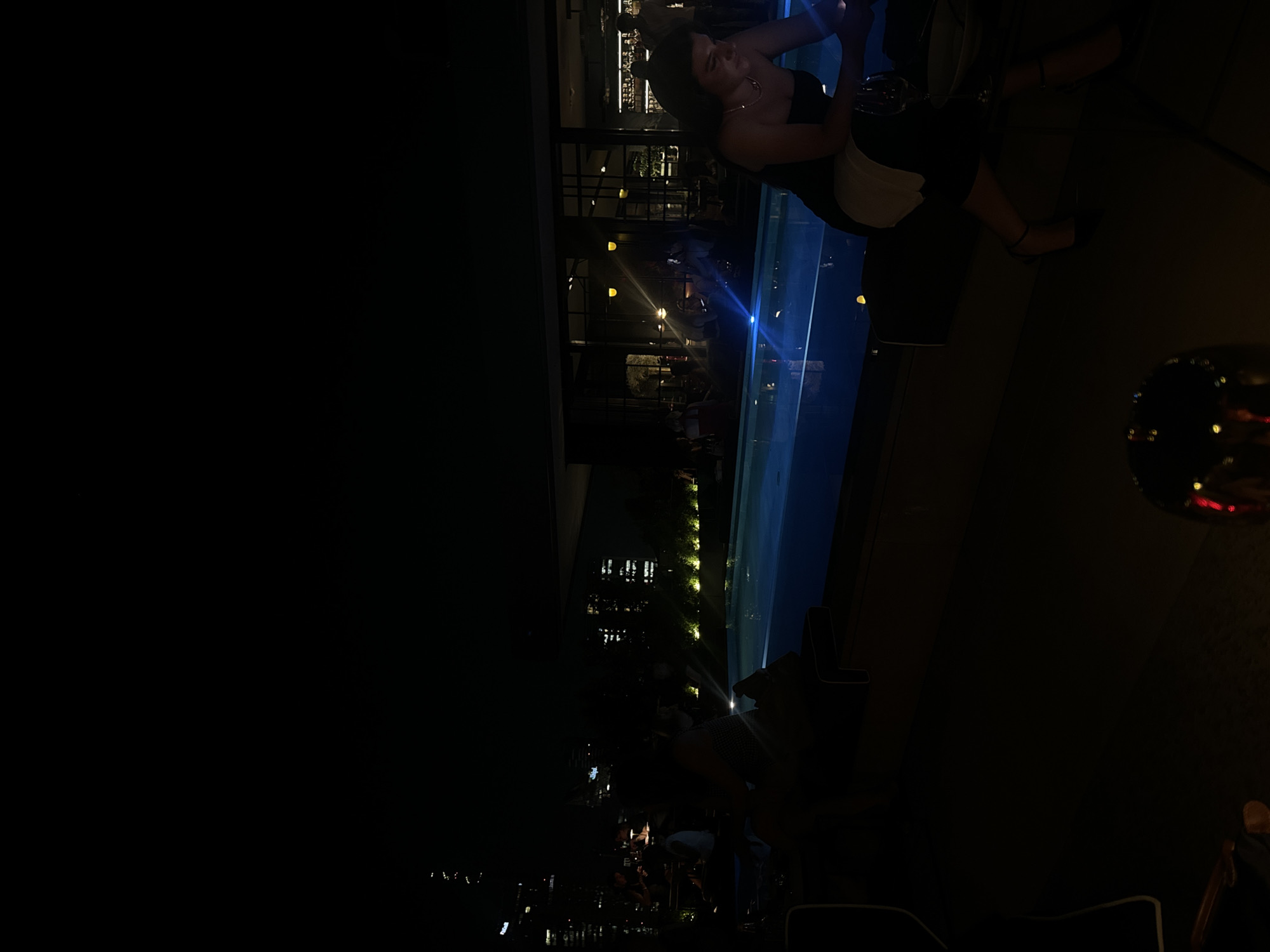 This image depicts a nighttime scene at a rooftop venue overlooking the Milan skyline, likely a restaurant or bar. A rectangular swimming pool with illuminated blue water is a central feature, reflecting the surrounding city lights. In the foreground, to the right, a woman in a black dress and high heels is seated, holding a glass of wine. Other patrons are visible in the background, seated at tables and near the pool. The urban landscape of Milan, with its illuminated skyscrapers, provides a dramatic backdrop to the scene. The lighting is dim, characteristic of a night setting, with the pool and building lights being the primary sources of illumination.